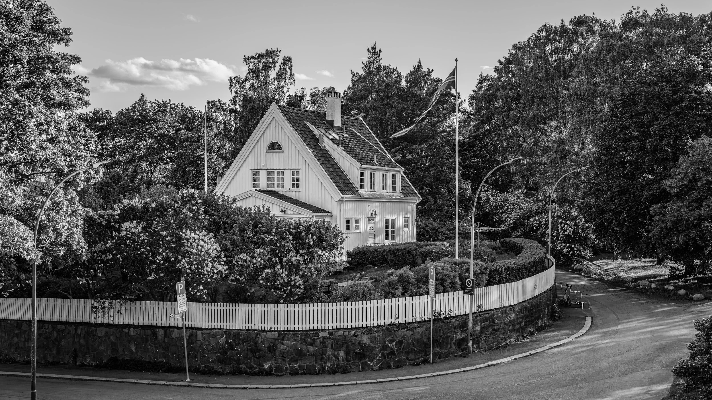 A residential house surrounded by trees and bushes, with a curved road and parking signs in the foreground, and flags on poles near the house.