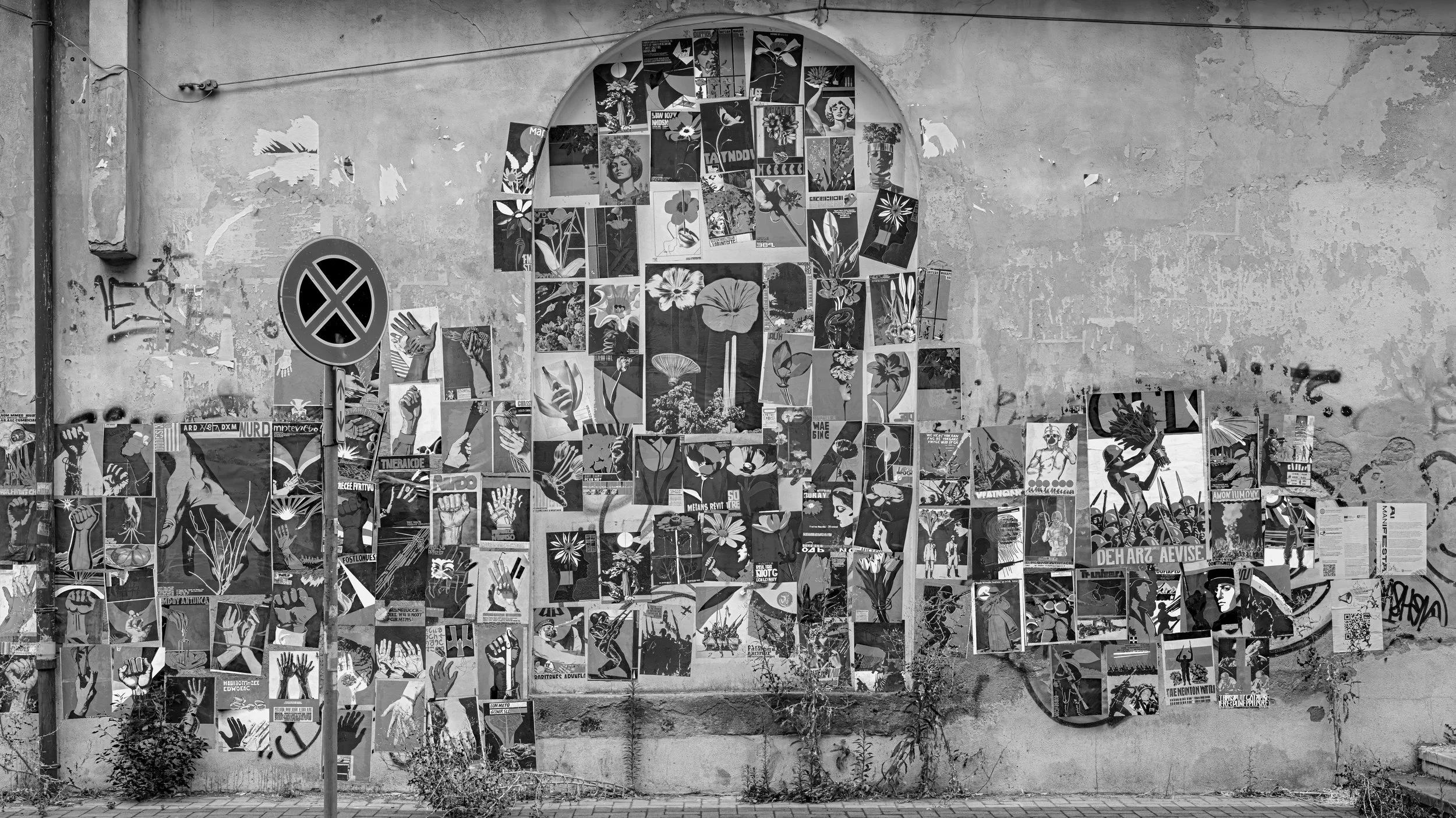 A black and white street mural composed of various posters and graffiti on a rough wall. The mural features artwork including flowers, faces, and protest images with fist symbols.