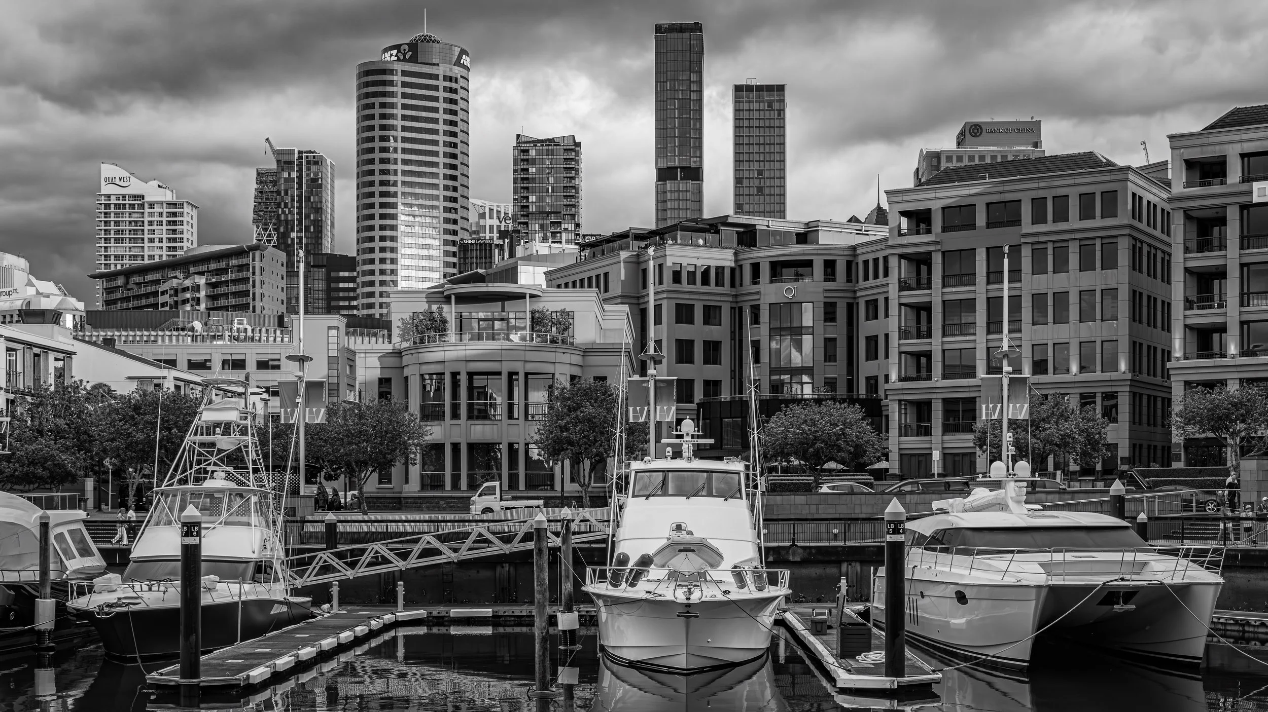 A marina with several boats docked in the foreground, and a city skyline with tall buildings under a cloudy sky in the background.