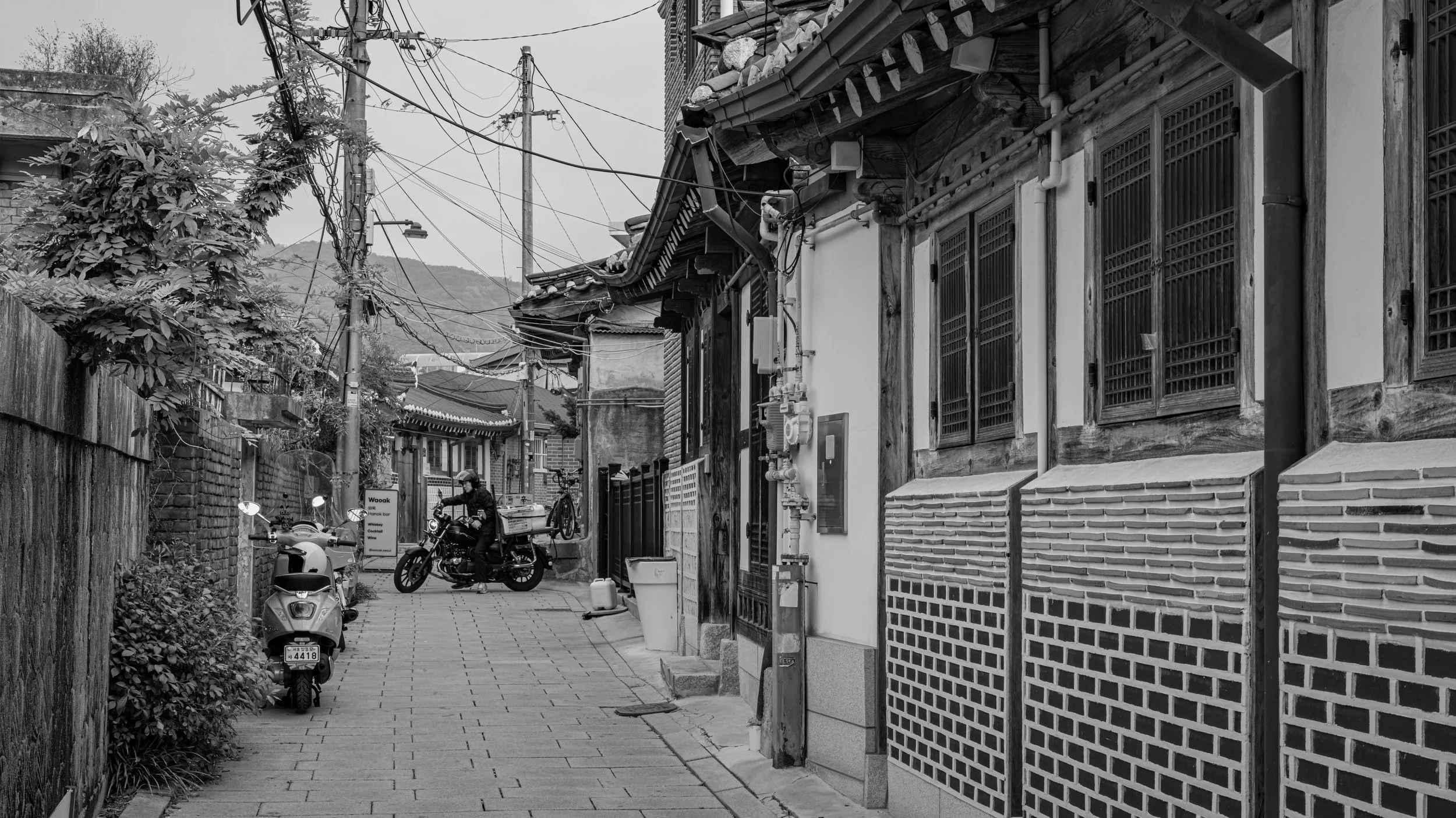 A black-and-white photo of a narrow alleyway with traditional buildings, electrical wires overhead, and motorcycles parked along the side. A person is riding a motorcycle in the distance.