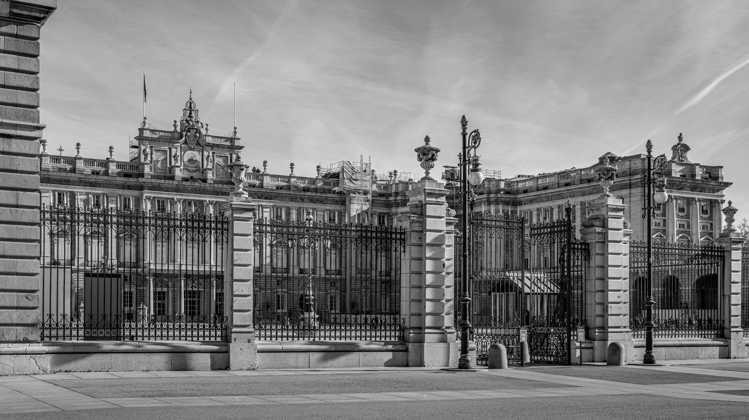 Black and white photo of an ornate historic building with detailed architecture, topped with sculptures and a clock, seen through a wrought iron fence with stone pillars and decorative lamp posts in the foreground.