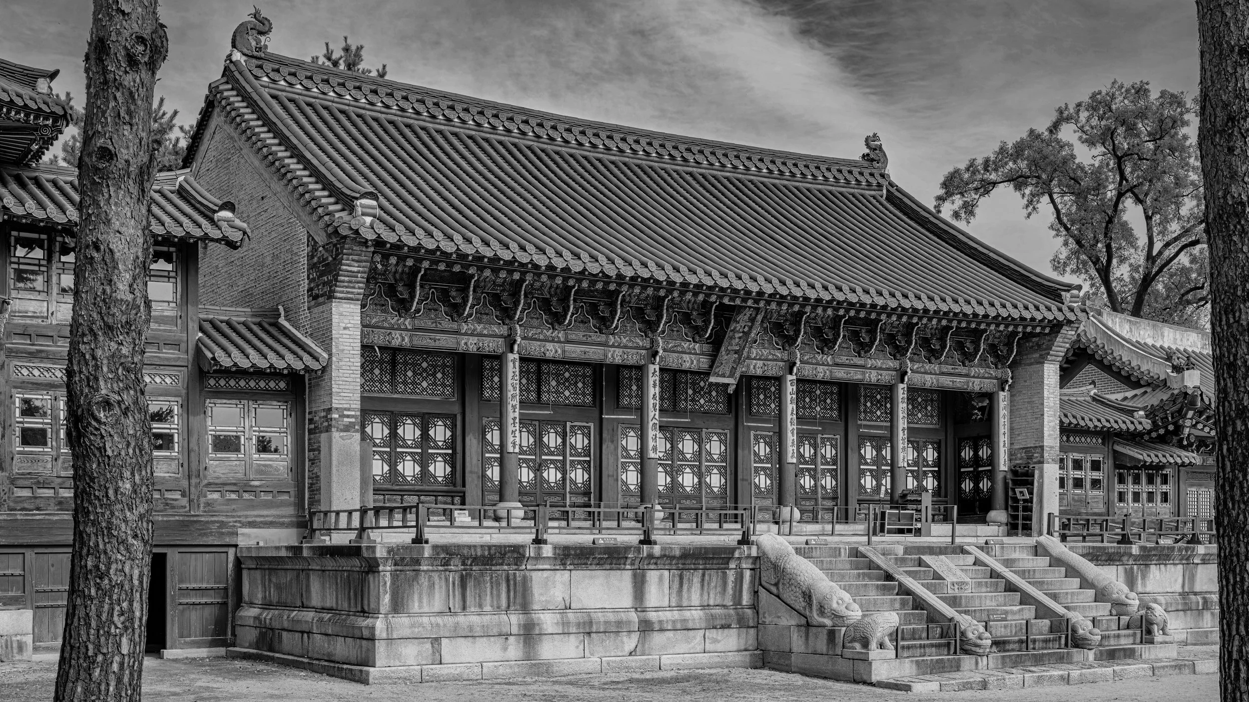 A traditional Asian temple with ornate architectural details, multiple tiled roofs, and stone dragon statues on stairs, framed by trees in black and white. Seoul, Korea.