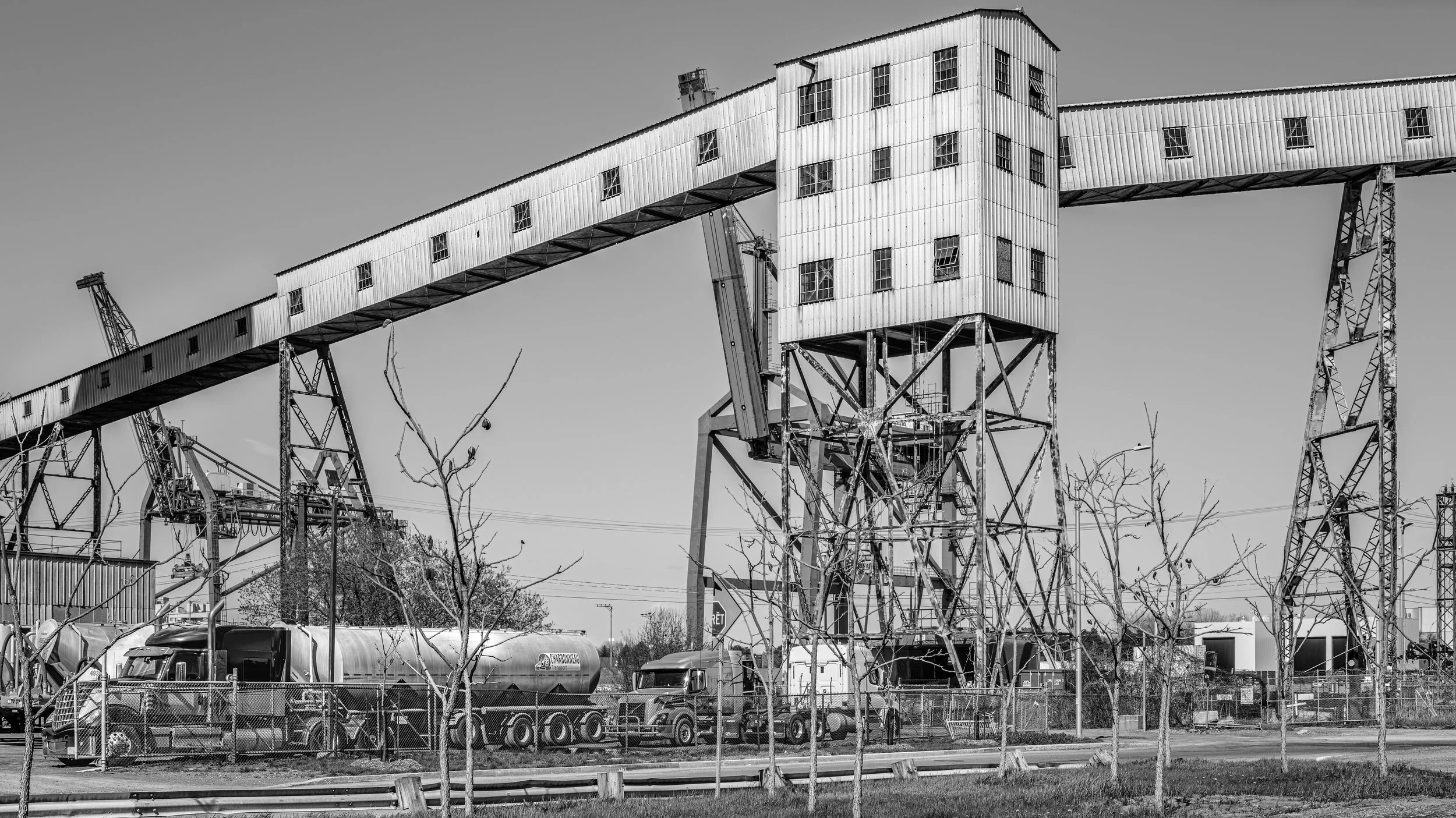 Black and white photo of an industrial landscape with a large metal building on stilts connected by a long elevated enclosed walkway. The structures appear old and weathered, with construction equipment and trucks below, and a parking lot with a car 