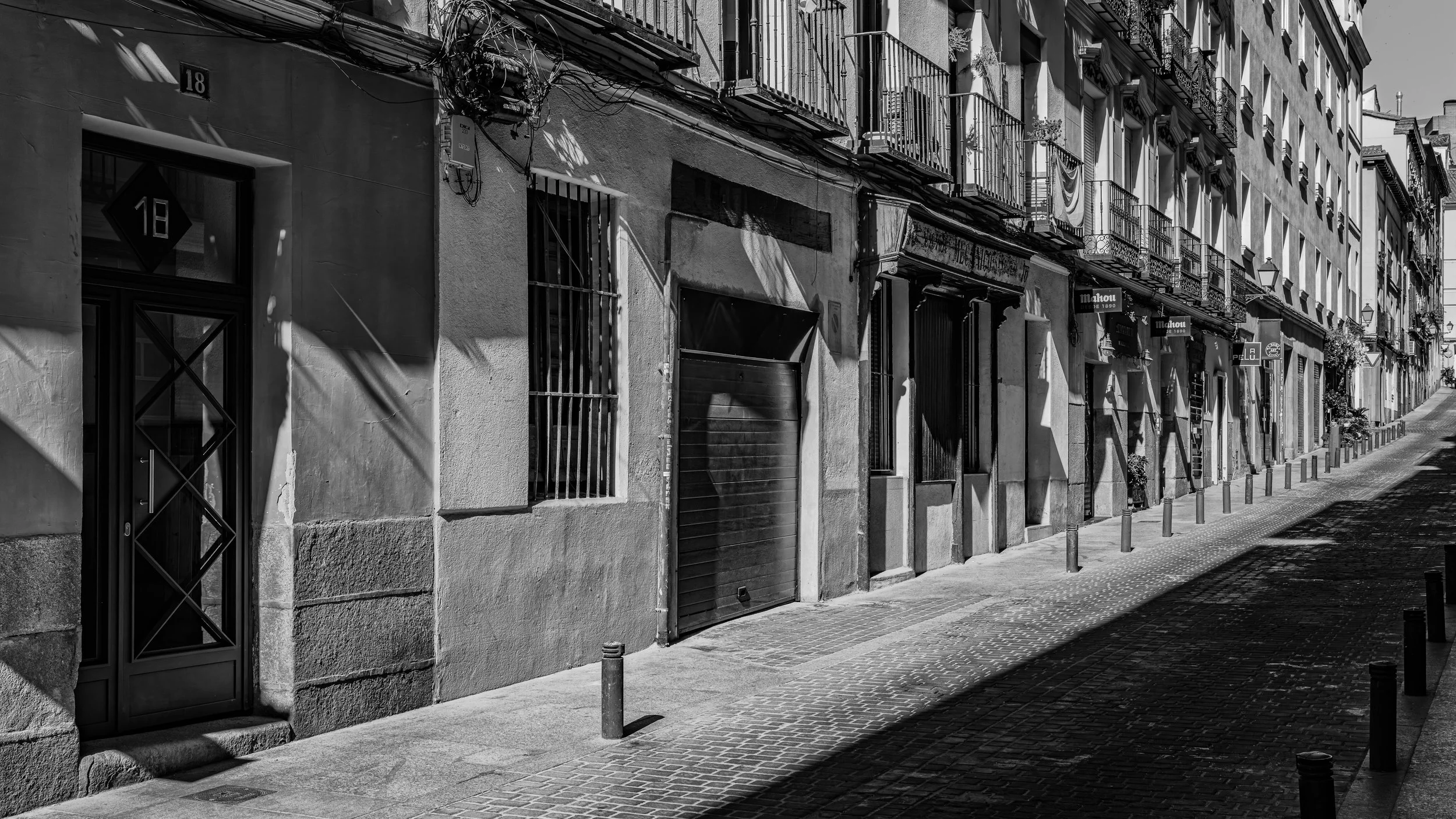 Black and white photo of a European street with row of apartment buildings with balconies, small shops, and a cobblestone sidewalk with posts. Shadows cast by the buildings.