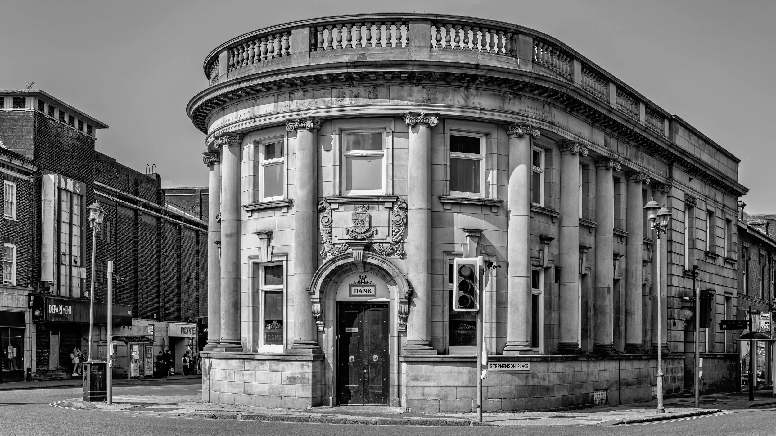 A black and white photo of a historic building with classical columns, decorative crest, and an arched entrance, located on a city street with street signs and lampposts. Chesterfield, Derbyshire, UK.