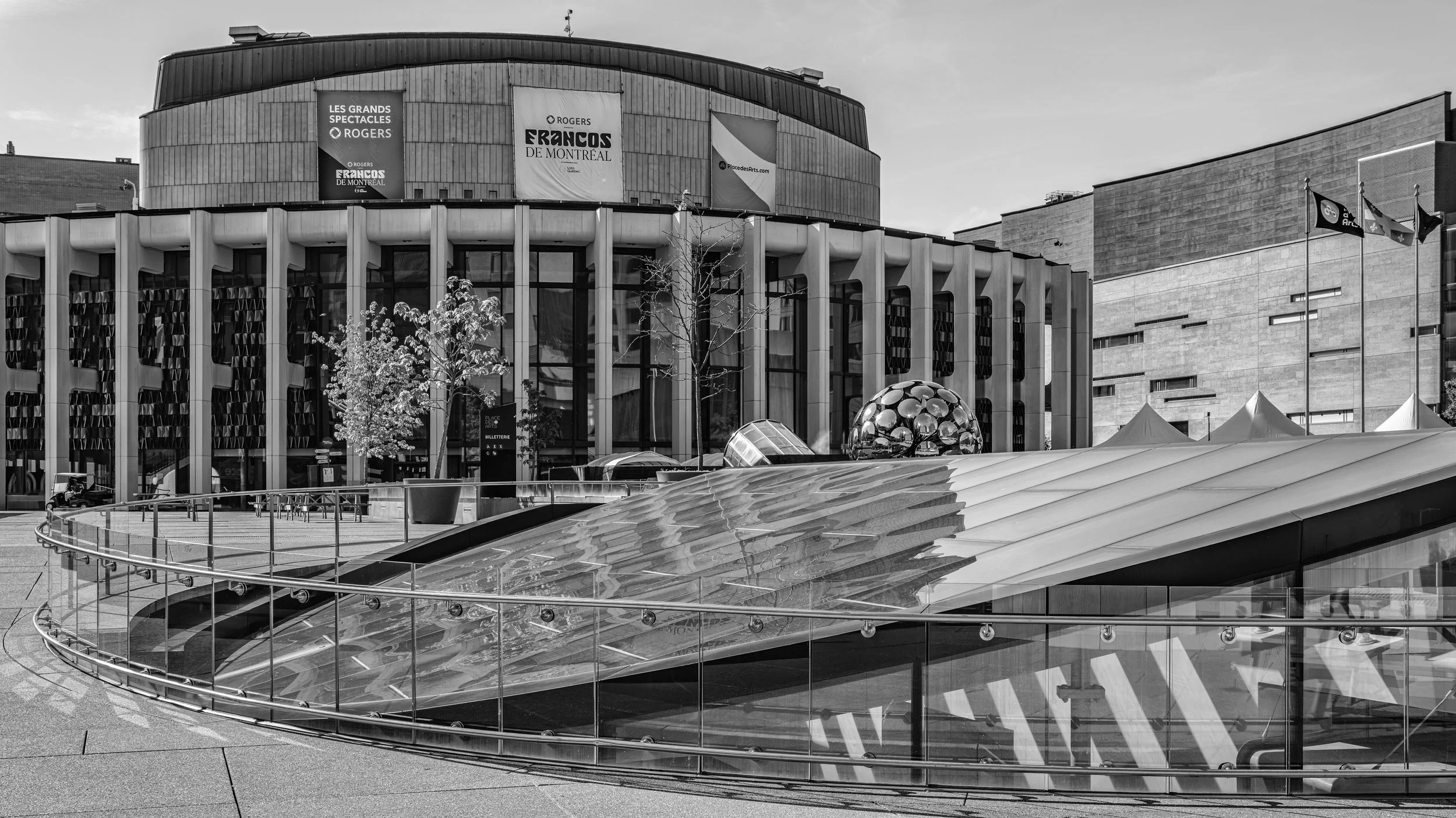 A modern building with banners advertising the Les Grands Spectacles at Place des Arts in Montreal, featuring a spherical mirror sculpture and a curved glass and metal awning in the foreground.