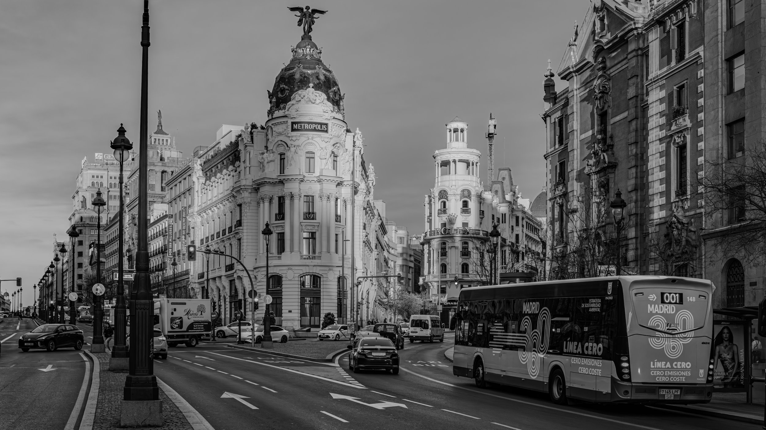 Black and white photo of a busy city street with cars and buses, historic buildings with ornate architecture, and a streetlamp in the foreground.