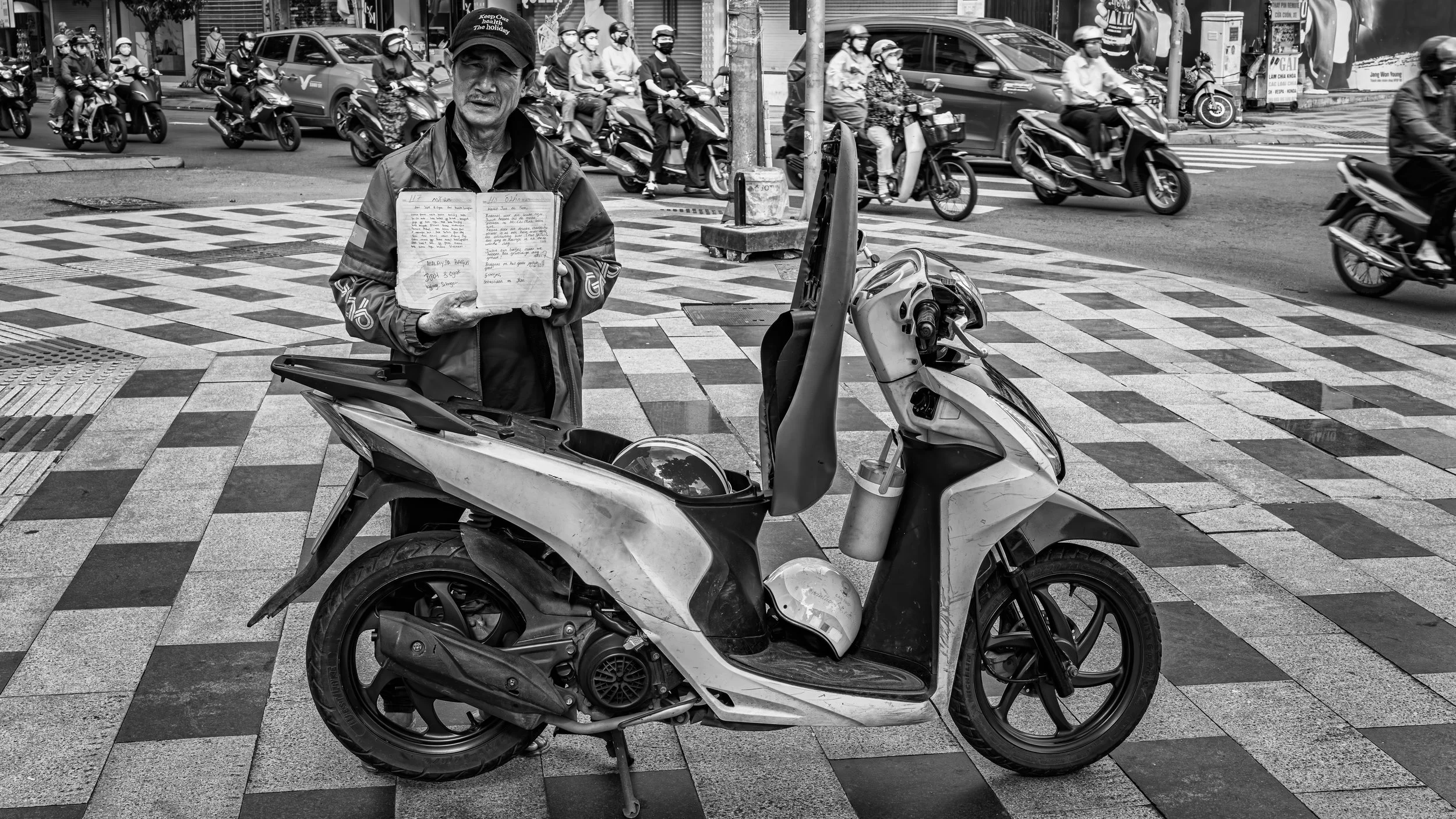 A man standing on the street holding a notebook, with motorcycles and cars passing by in the background. The scene is captured in black and white.