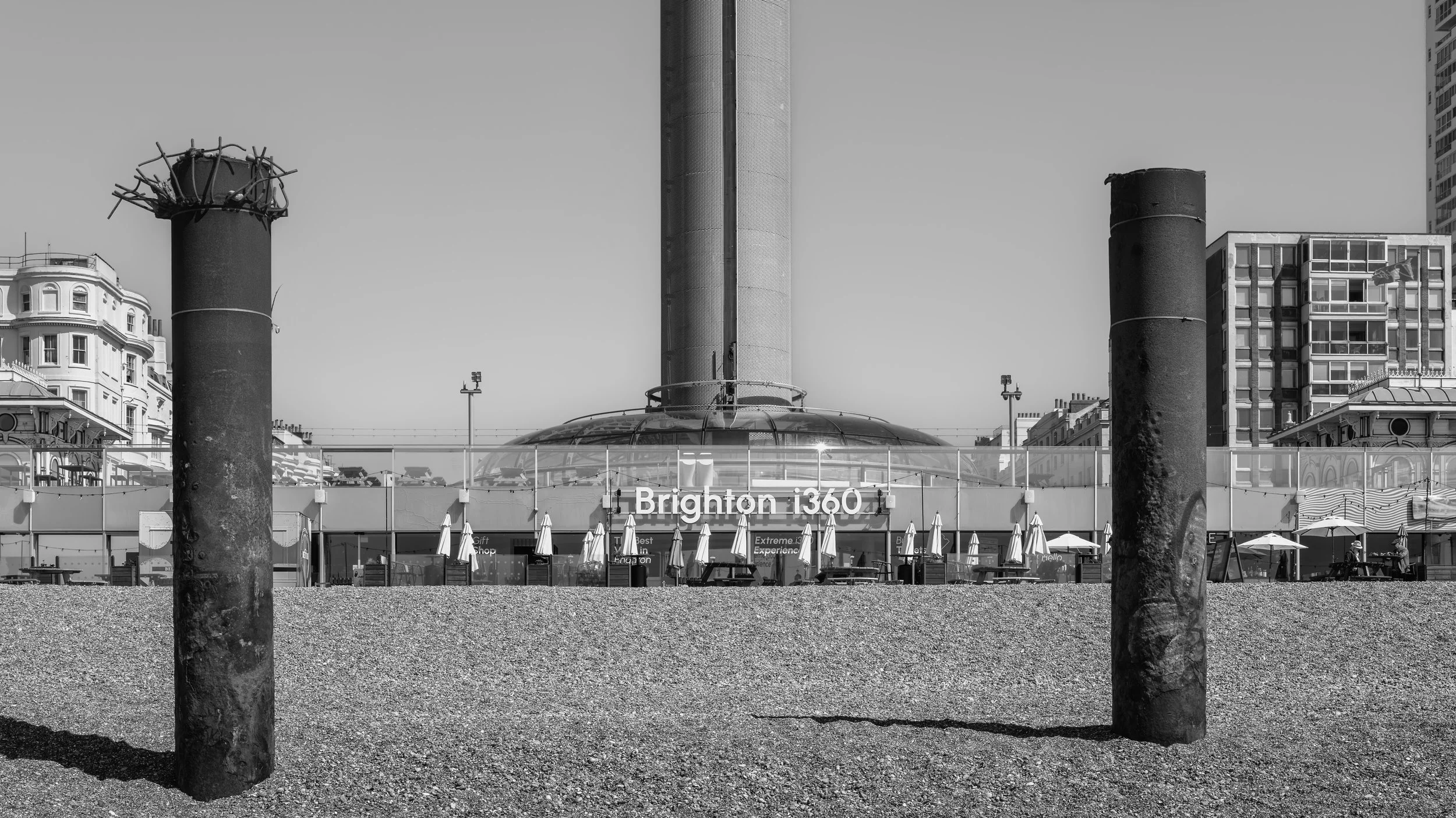Black and white photo of Brighton i360 tourist attraction with two large metal chimneys in front, beach area, and buildings in the background.