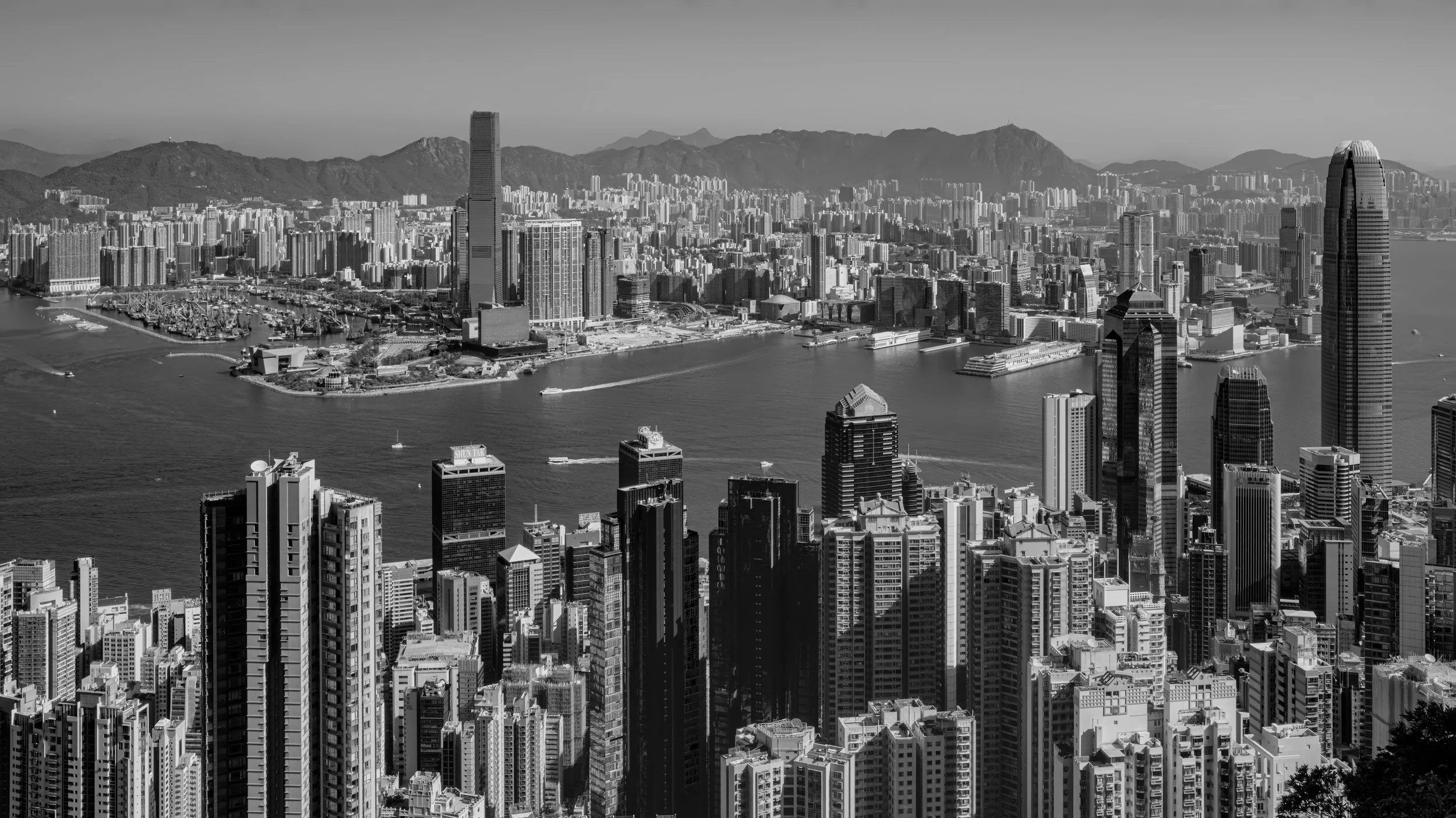 Black and white aerial view of a densely populated cityscape with tall skyscrapers, a harbor with boats, and mountain range in the background.