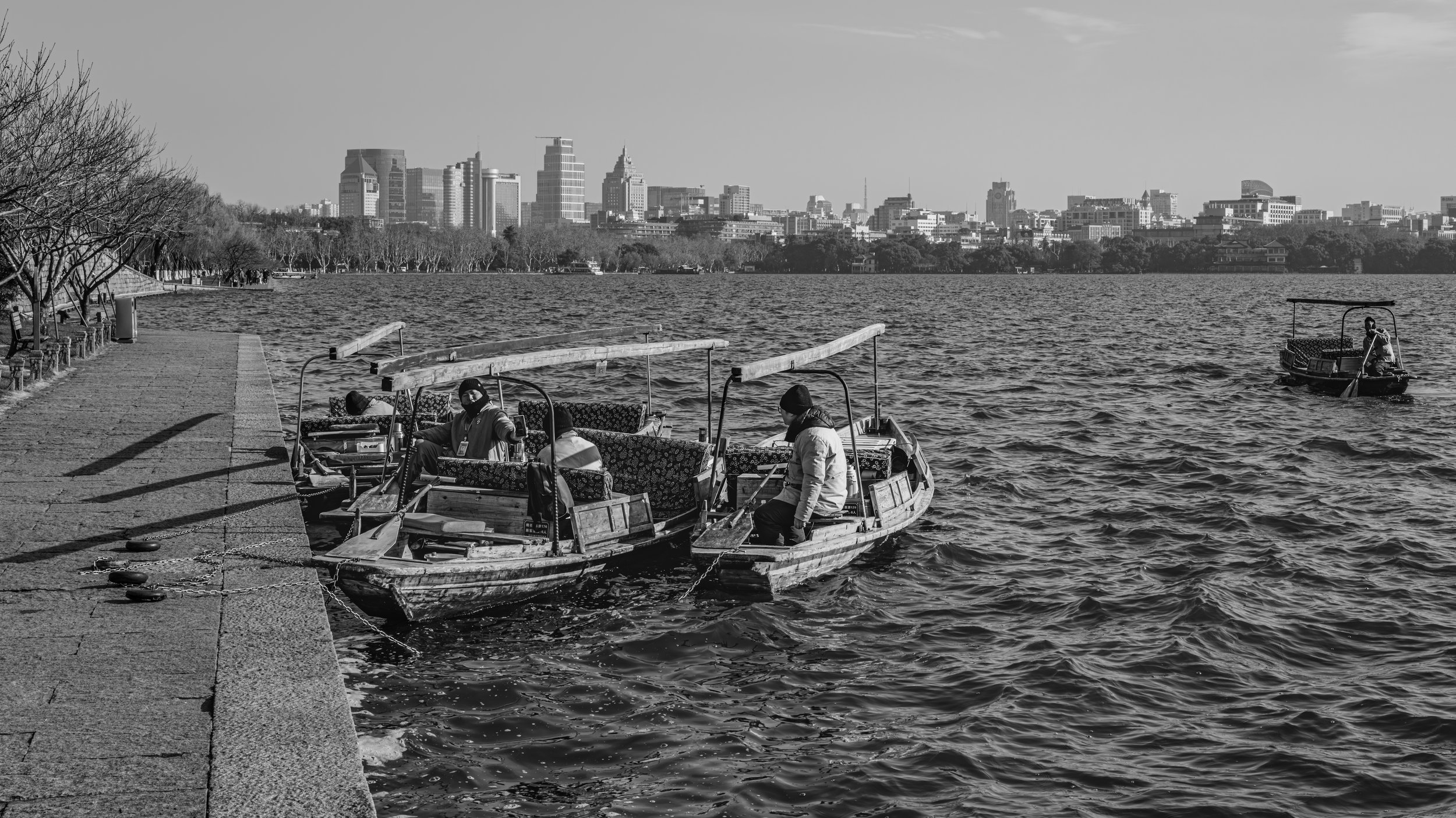 Black and white photo of a city skyline across a lake with boats and people on the water, and leafless trees along the shoreline.