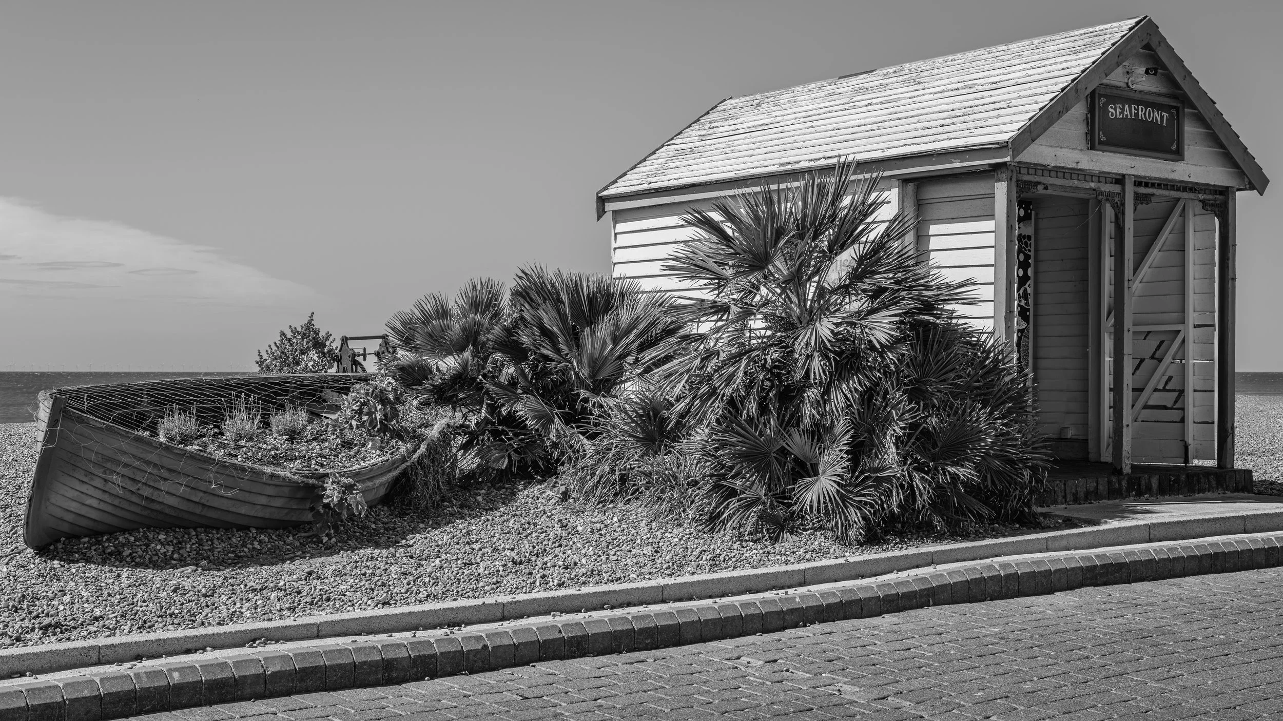 A black and white photo of a small wooden seaside structure labeled 'SEAFRONT'. Beside it, a boat is tipped over on a gravelly area with plants growing inside it. In the background, there is a body of water and sky.