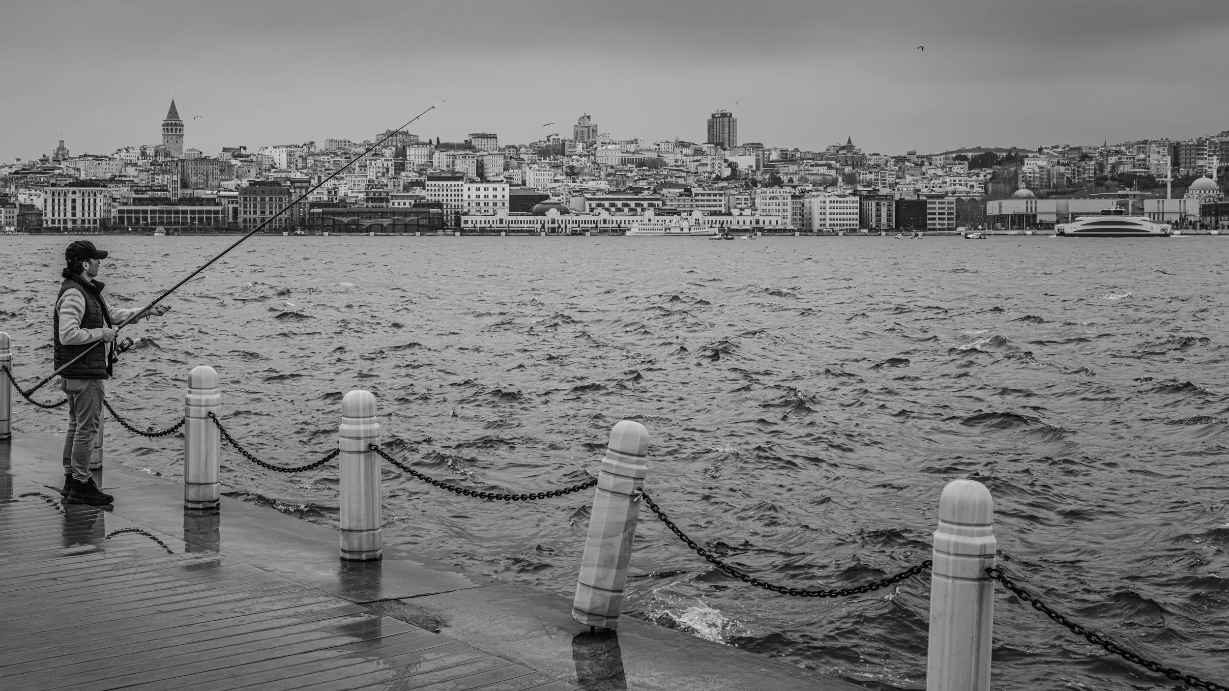 A person fishing off a dock by the water with a city skyline in the background in black and white.