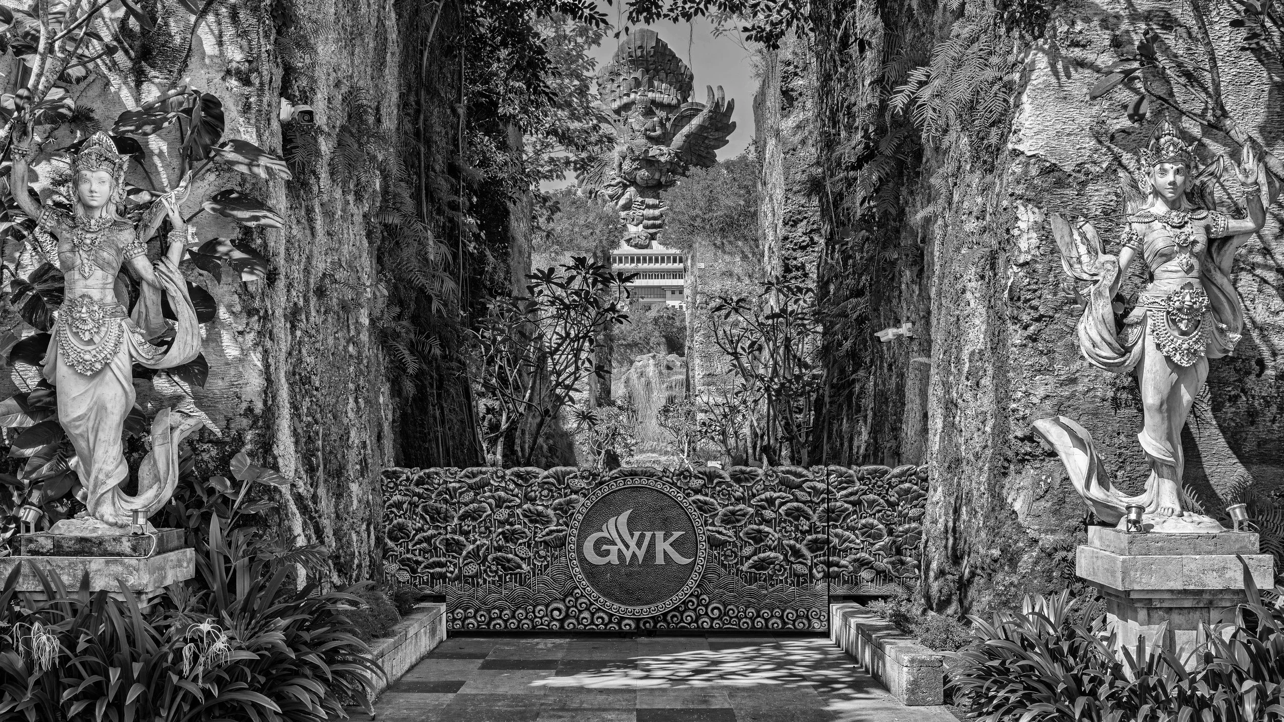 Black and white photo of a decorative temple entrance with two ornate female statues on either side, surrounded by plants and rocks, with a central gate displaying the initials 'GWK' and a carved circular emblem below.