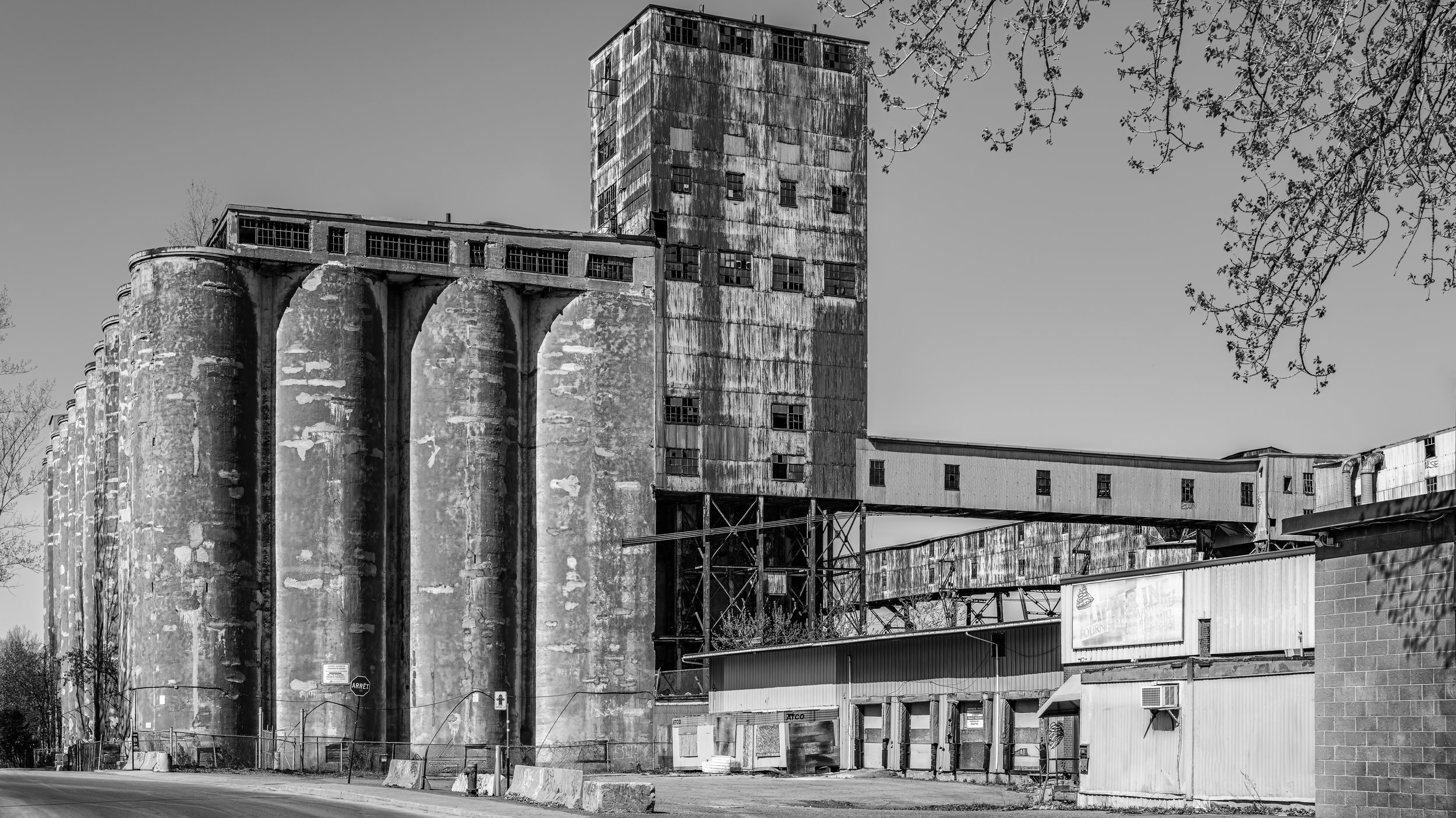 Black and white photo of an old, abandoned industrial building with tall silos and a large tower, surrounded by smaller structures and trees.