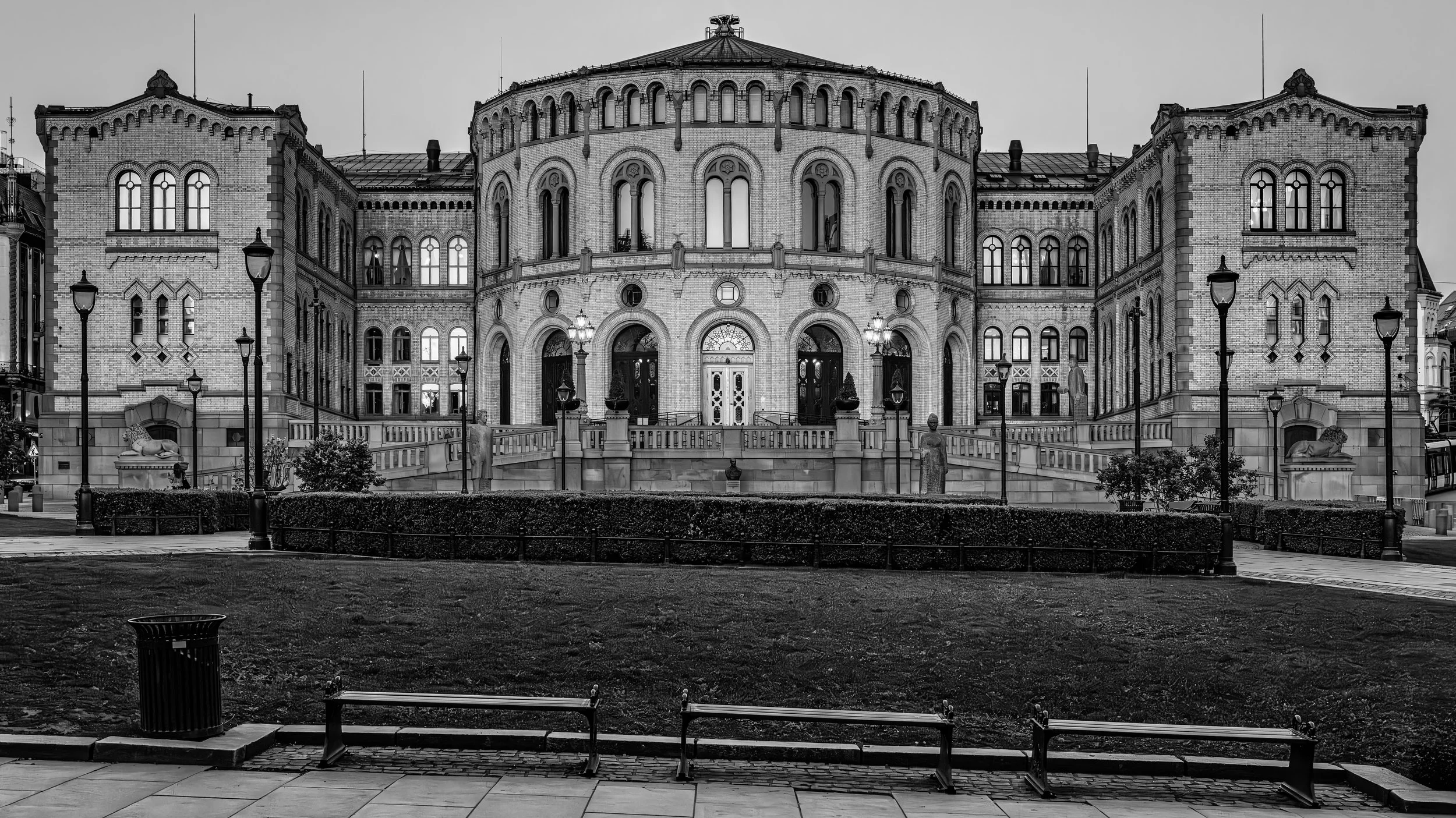 A large historic building with a rounded central section and two symmetrical wings, constructed with brick and decorative stone accents, viewed in black and white. The building features tall arched windows, lantern-style streetlights, and stone sculp