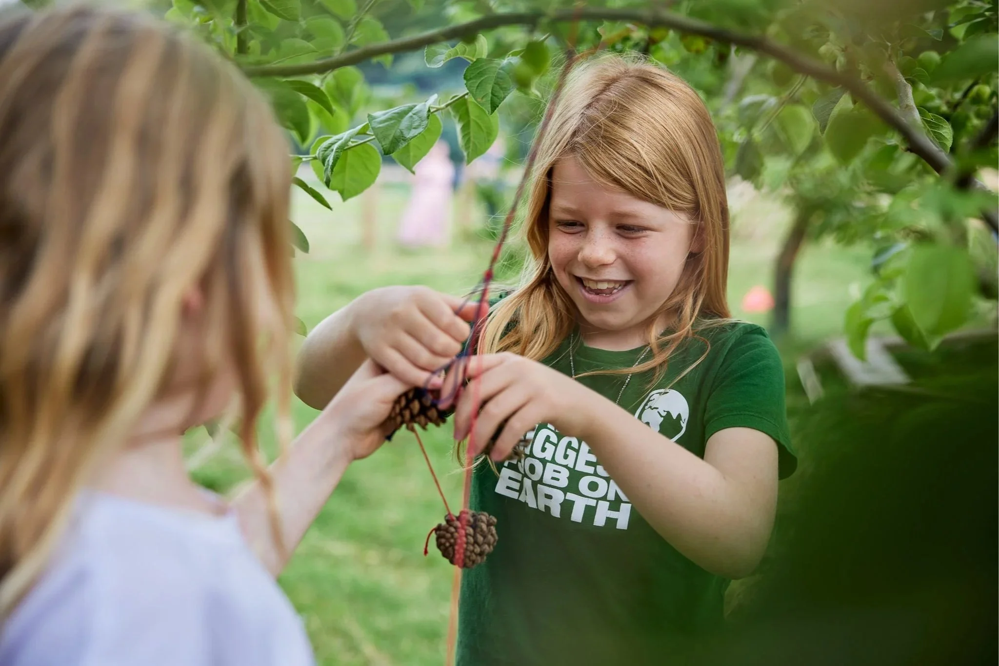 Two children engaged in outdoor activity, making crafts with pinecones and string, surrounded by tree foliage.
