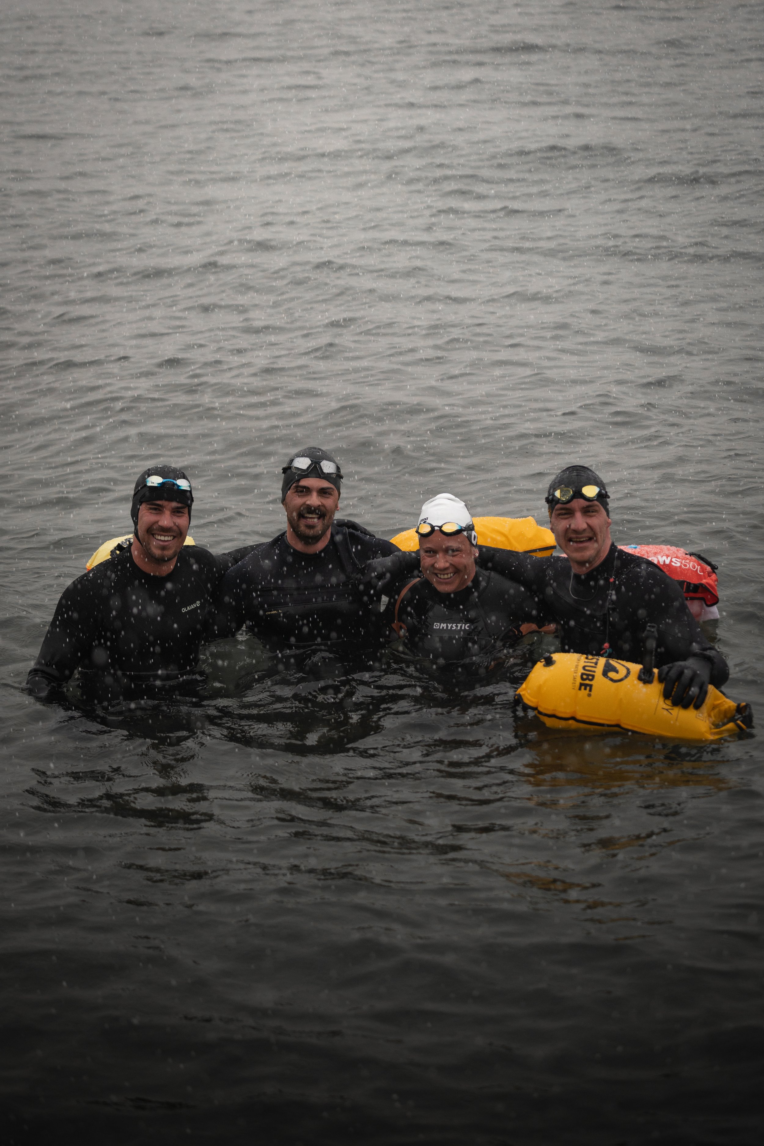 Vier Leute in Neoprenanzügen schwimmen lächelnd in einem Gewässer, einige tragen Schwimmbrillen. Zwei von ihnen halten gelbe Schwimmbojen.