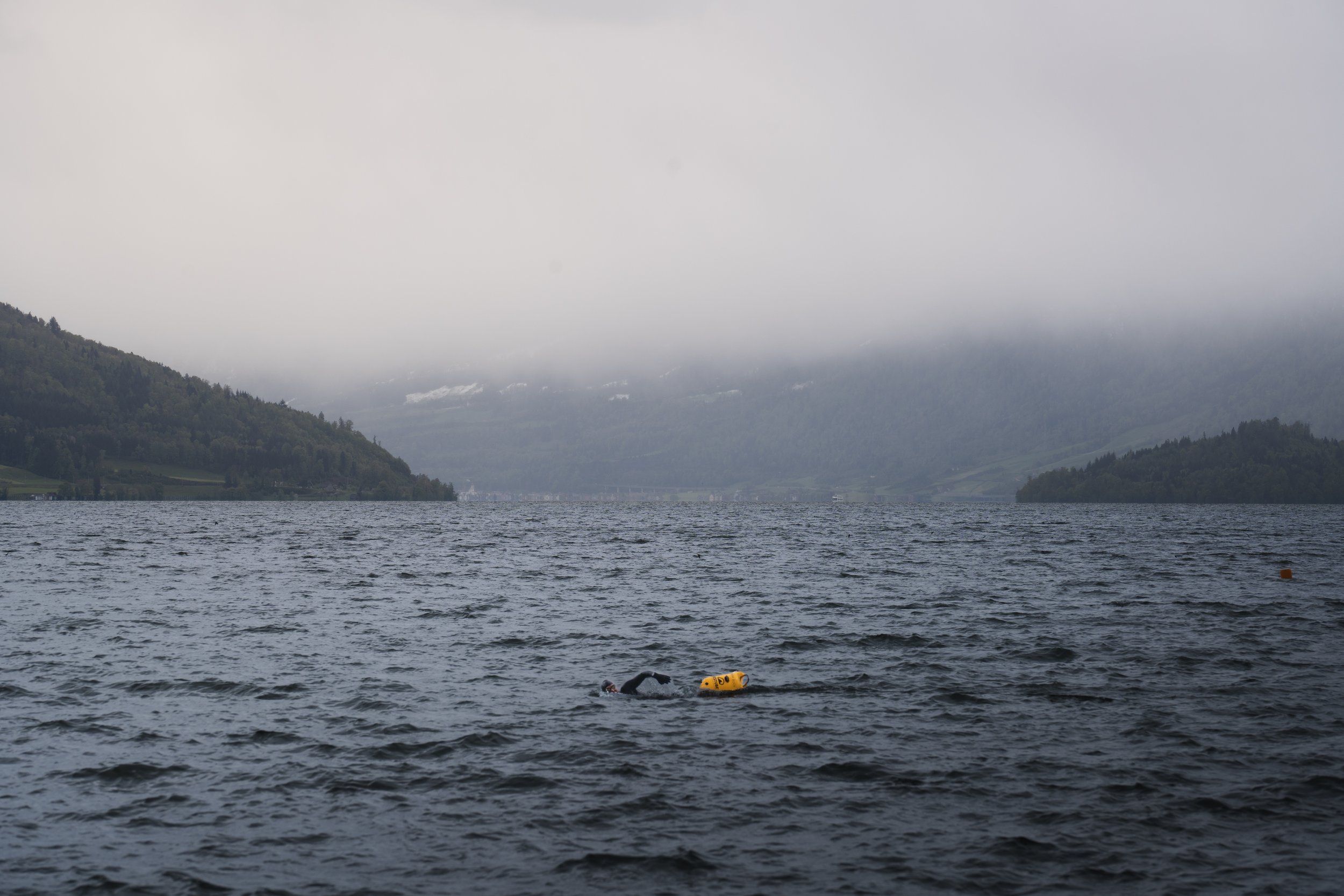 Ein Schwimmer mit gelber Boje im Wasser eines großen Sees, umgeben von bewaldeten Hügeln und dichtem Nebel.