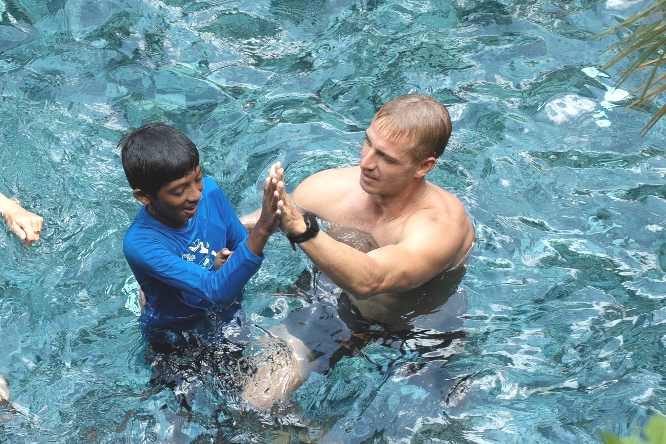 Zwei Personen im Wasser, ein Mann hebt ein Kind hoch. Der Mann ist muskulös und trägt keine Shirts, während das Kind ein blaues Langarmshirt trägt. Beide lachen und scheinen Spaß zu haben.