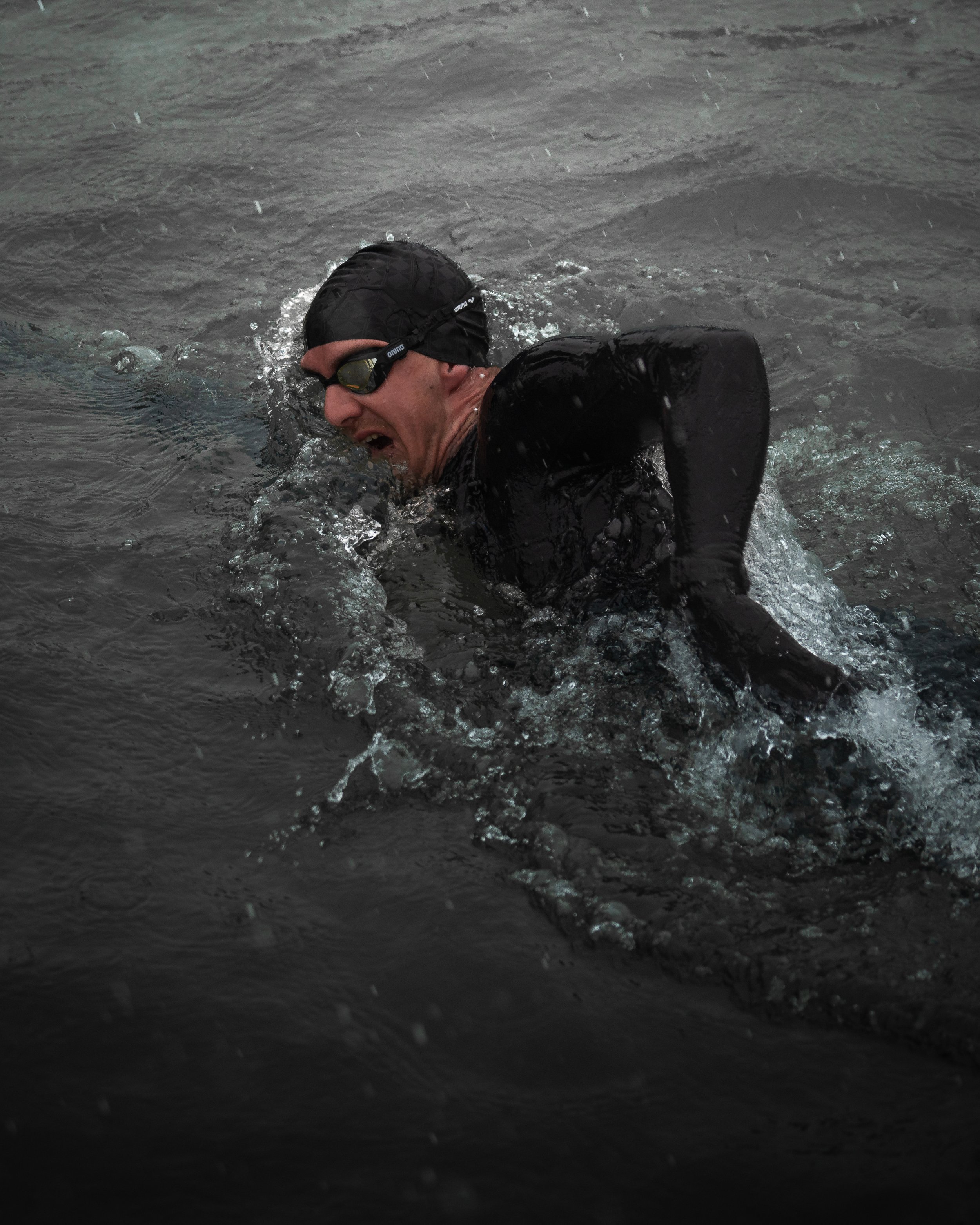 Ein Schwimmer in einem Neoprenanzug und einer schwarzen Badekappe schwimmt im Wasser. Er trägt eine Schwimmbrille und bewegt sich durch die Wellen.
