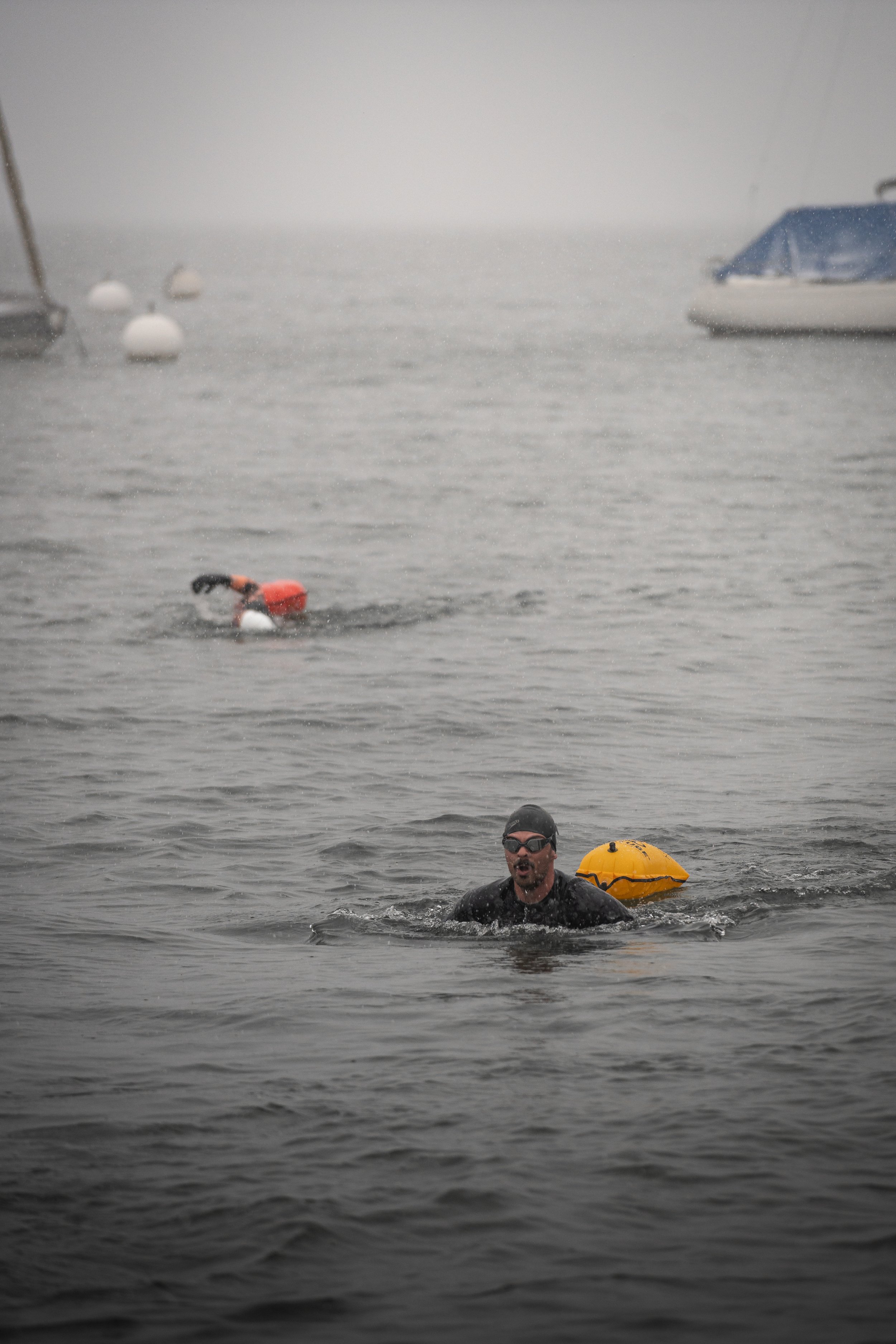 Zwei Schwimmer im offenen Wasser mit Schwimmbojen im Hintergrund, trübes Wetter, Boote im Wasser.