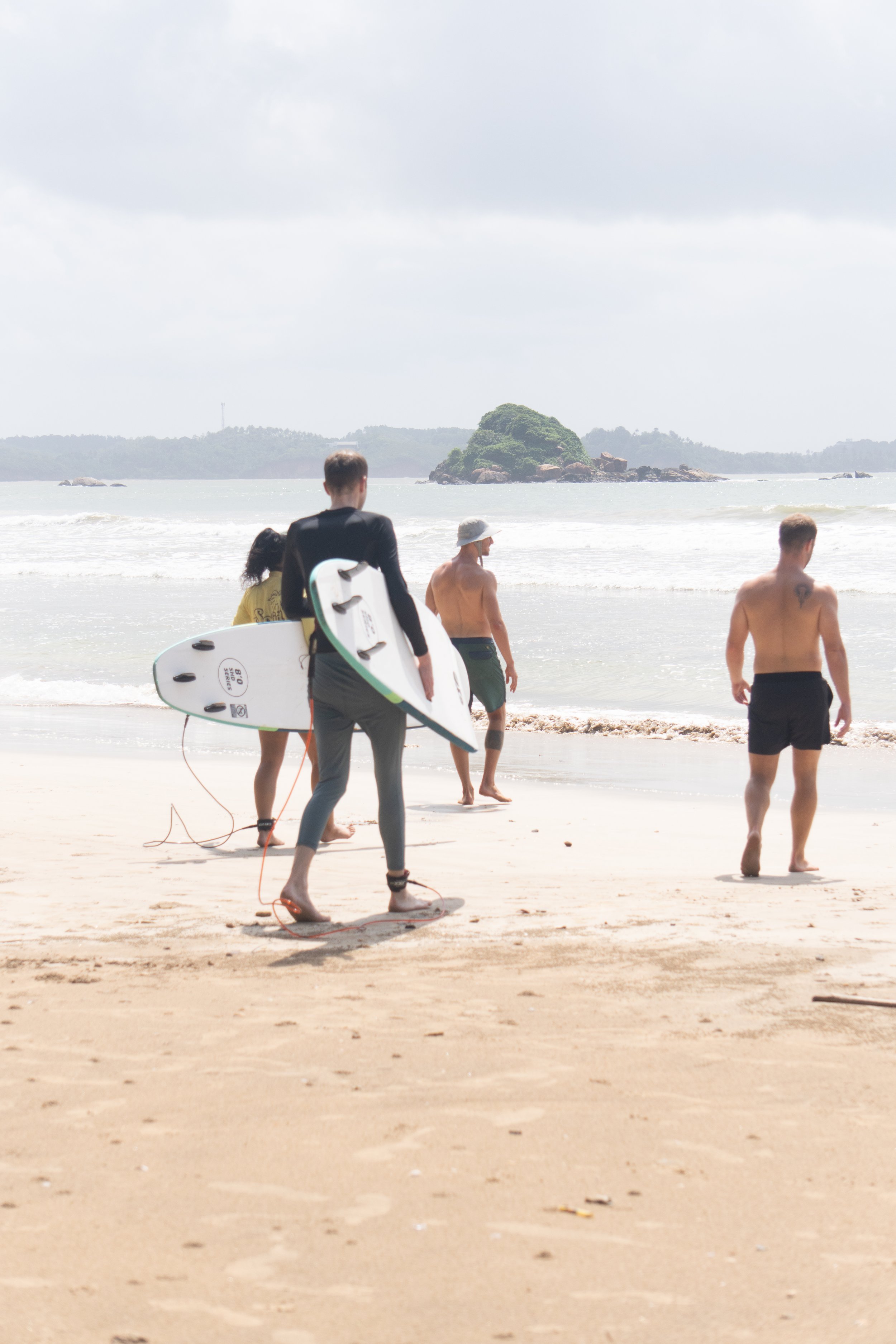 Gruppe von Menschen mit Surfboards am Strand, bereit zum Surfen; Meer und kleine Insel im Hintergrund.