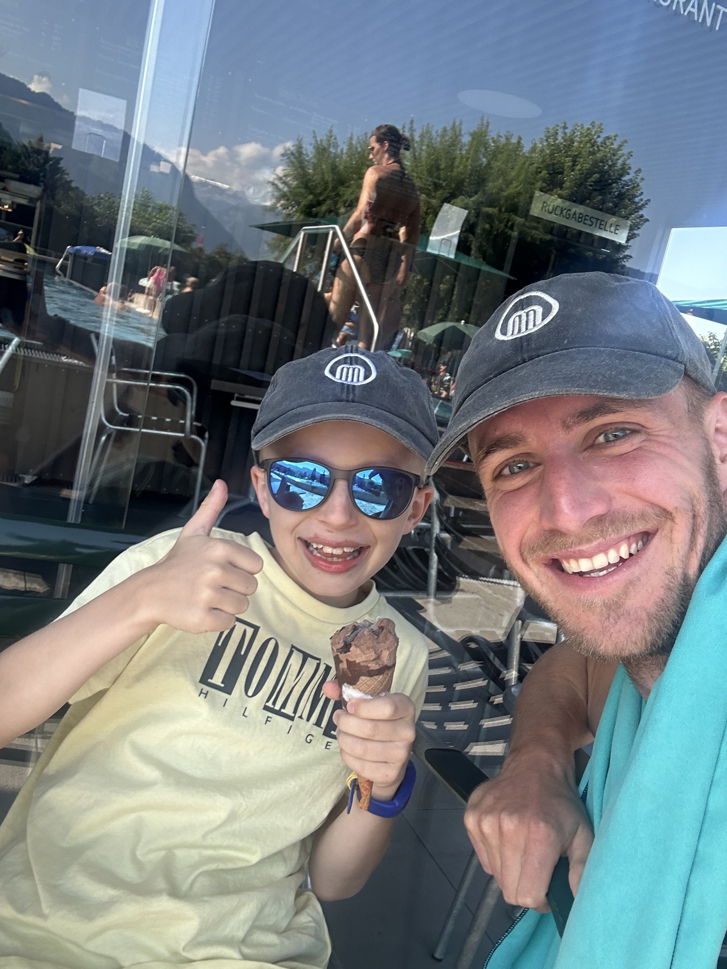 Zwei Personen mit Baseballmützen lächeln in die Kamera. Eine Person hält ein Schokoladeneis und zeigt einen Daumen hoch. Im Hintergrund reflektiert das Glas eines Gebäudes den Außenbereich mit Bäumen und Menschen in einem Pool.