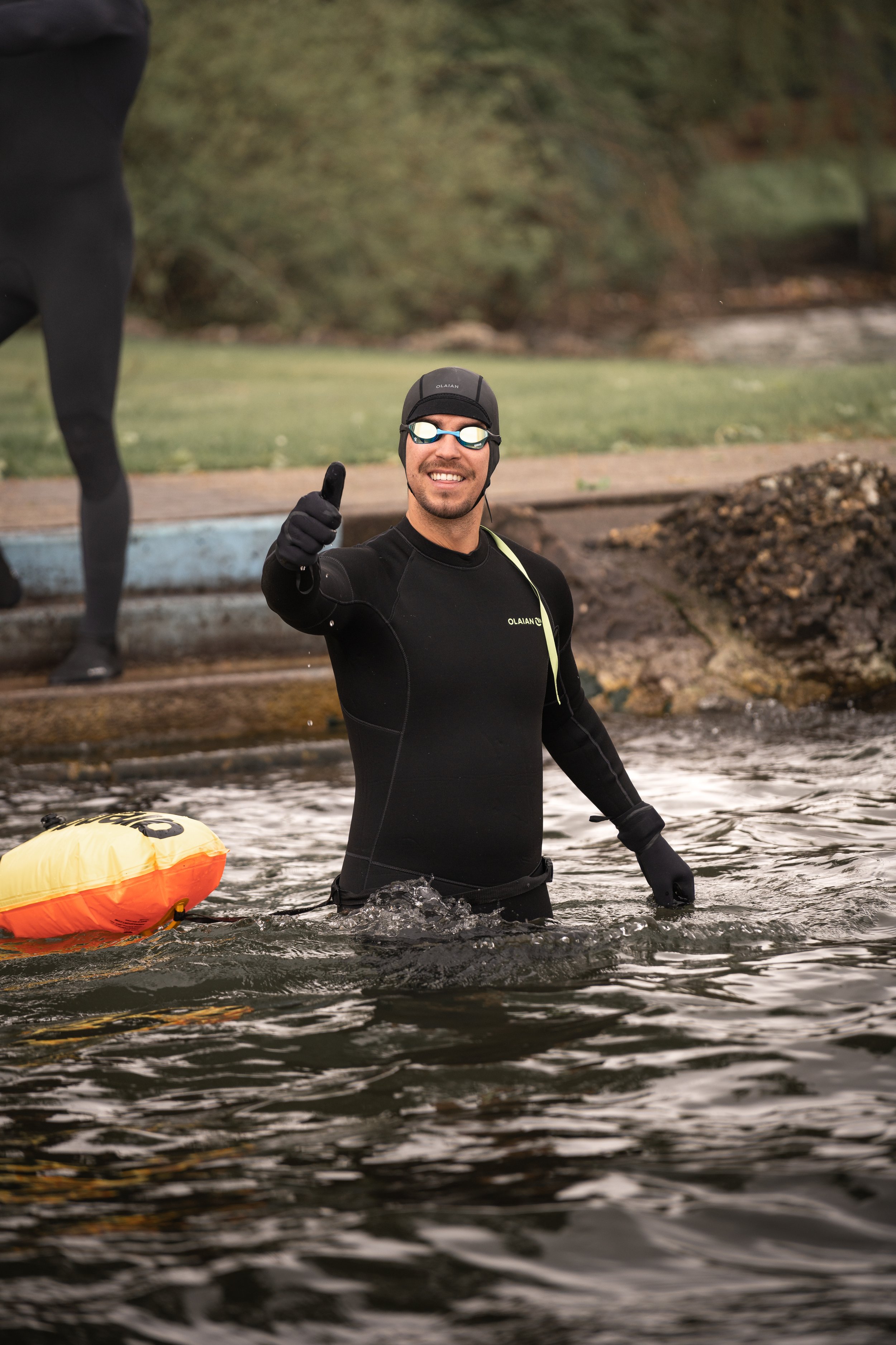 Ein Mann in einem Neoprenanzug steht im Wasser und zeigt mit einem Daumen nach oben. Er trägt eine Schwimmbrille und eine Schwimmkappe. Im Hintergrund ist ein weiterer Taucher zu sehen.