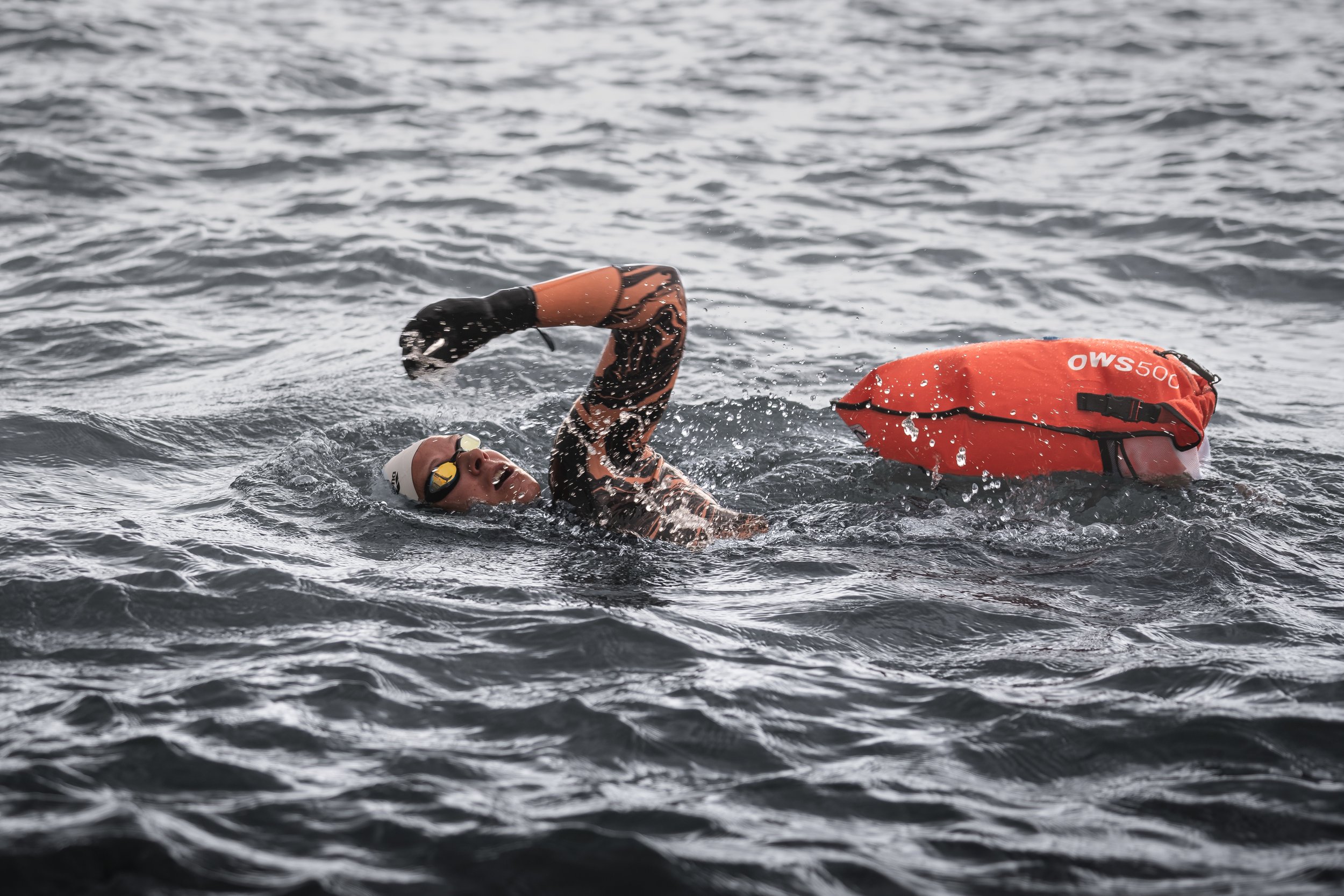 Ein Schwimmer mit Neoprenanzug, Schwimmbrille und Badekappe schwimmt im offenen Wasser mit einer orangefarbenen Schwimmboje.