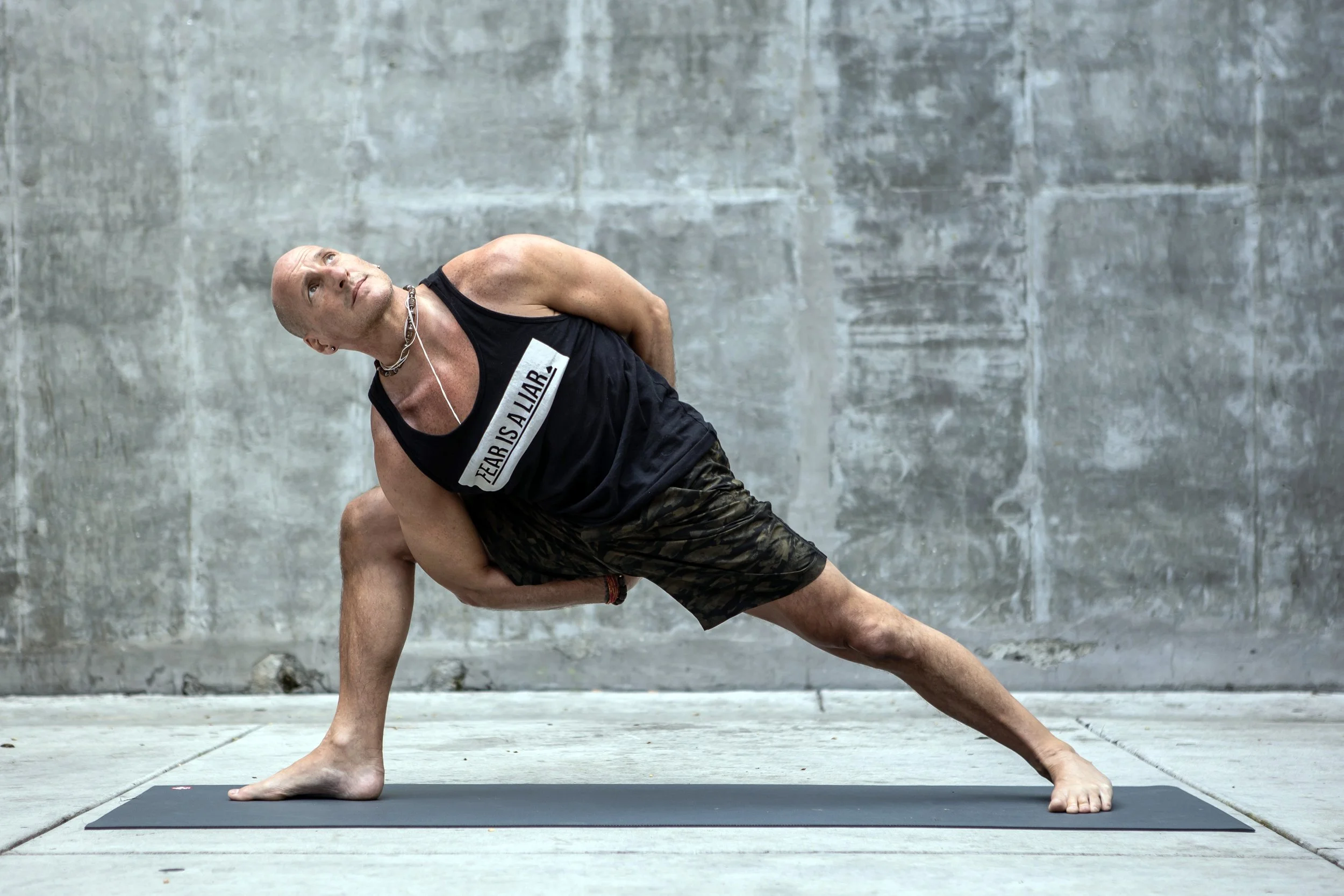 A man performing a yoga pose on a black yoga mat against a concrete wall, wearing a black tank top with 'TEARS IS A LIE.' written on it and camouflage shorts.
