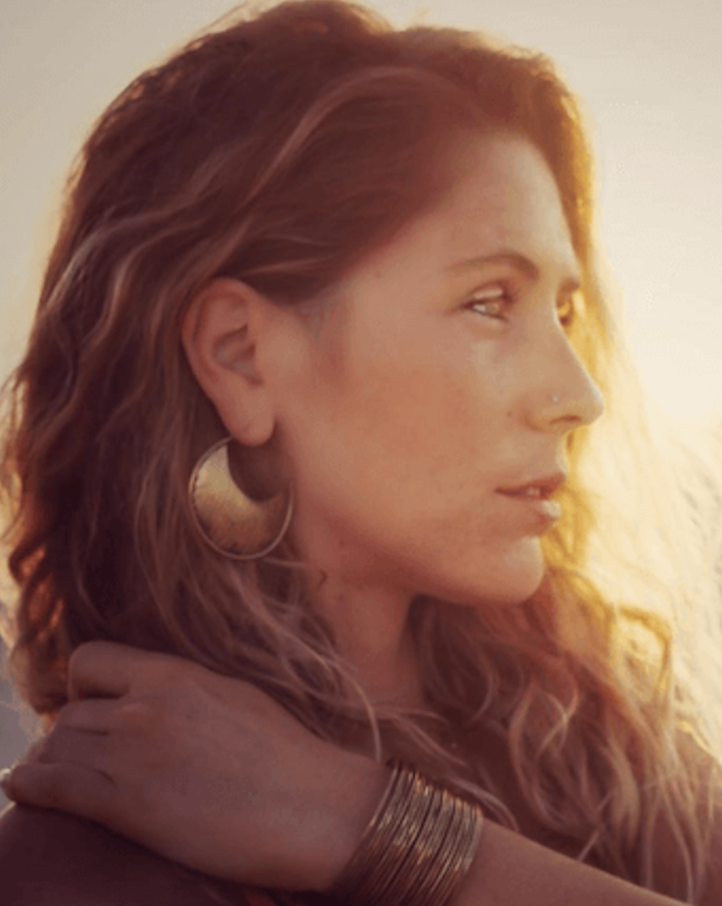 A woman with curly red hair and gold jewelry, looking to the right with a serene expression, in warm sunlight.