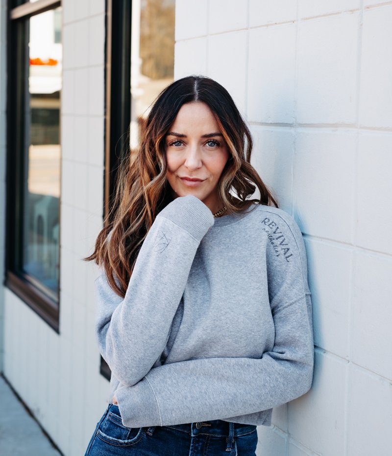Woman with wavy brown hair and blue eyes wearing a gray sweatshirt and jeans, leaning against a white brick wall outside a building with large windows.
