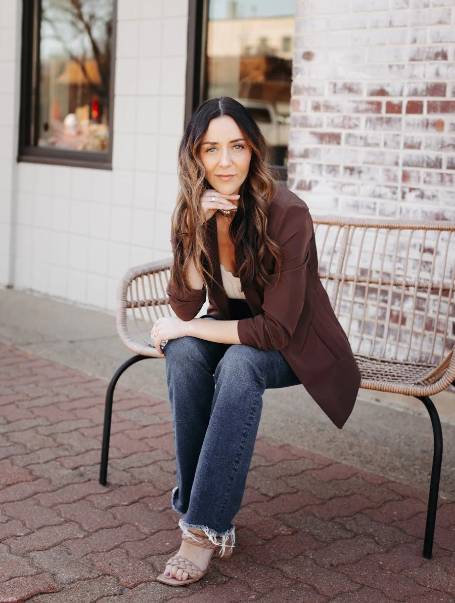A woman with long wavy brown hair sitting on a bench outside a building with brick and white tiled walls, looking at the camera with her chin resting on her hand.