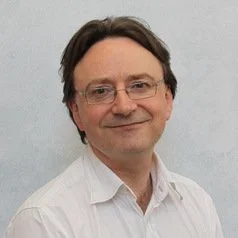 A man with glasses and brown hair wearing a white collared shirt, smiling against a plain light background.