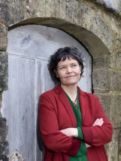 Close-up of Kate Raworth with short, dark, wavy hair wearing a green sweater and a red and white necklace, standing in front of a light-colored brick wall.