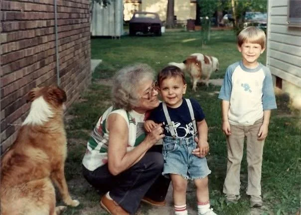 A woman with glasses kneeling next to a young girl and a boy, all smiling outdoors in a backyard with grass and dogs.