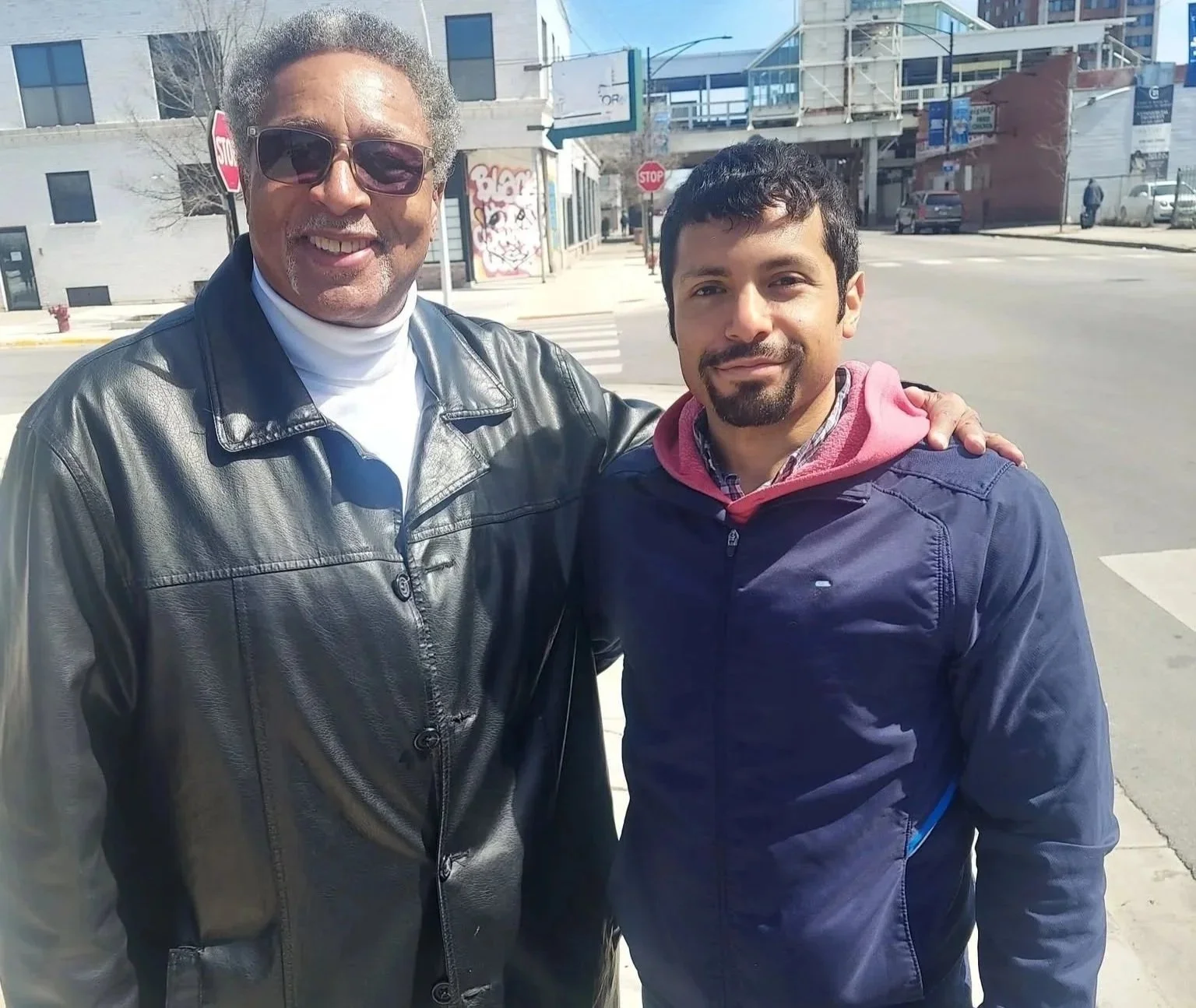 Two men standing outside on a city street, smiling at the camera. One man is wearing sunglasses and a leather jacket, the other has a beard and is wearing a navy jacket and hoodie.