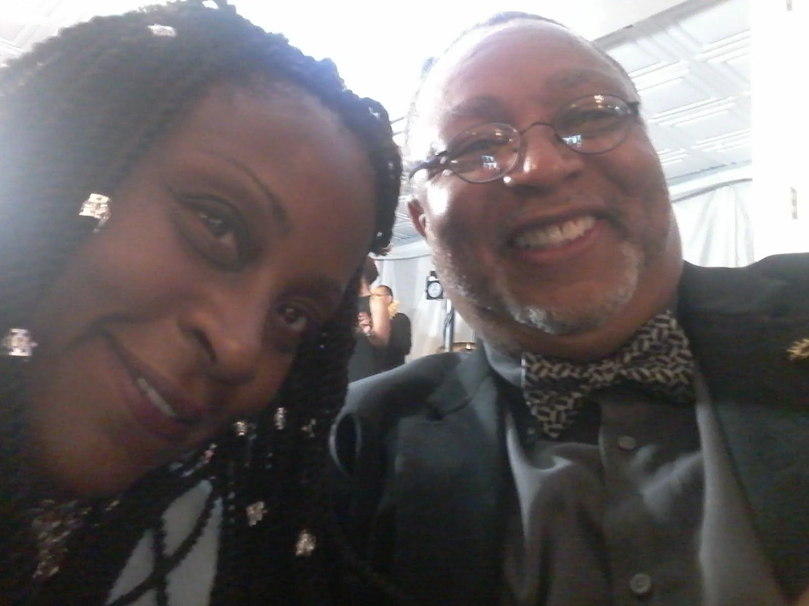 Two people taking a selfie at an indoor event, smiling. One person is a woman with braided hair, the other is an older man with glasses and a bow tie.