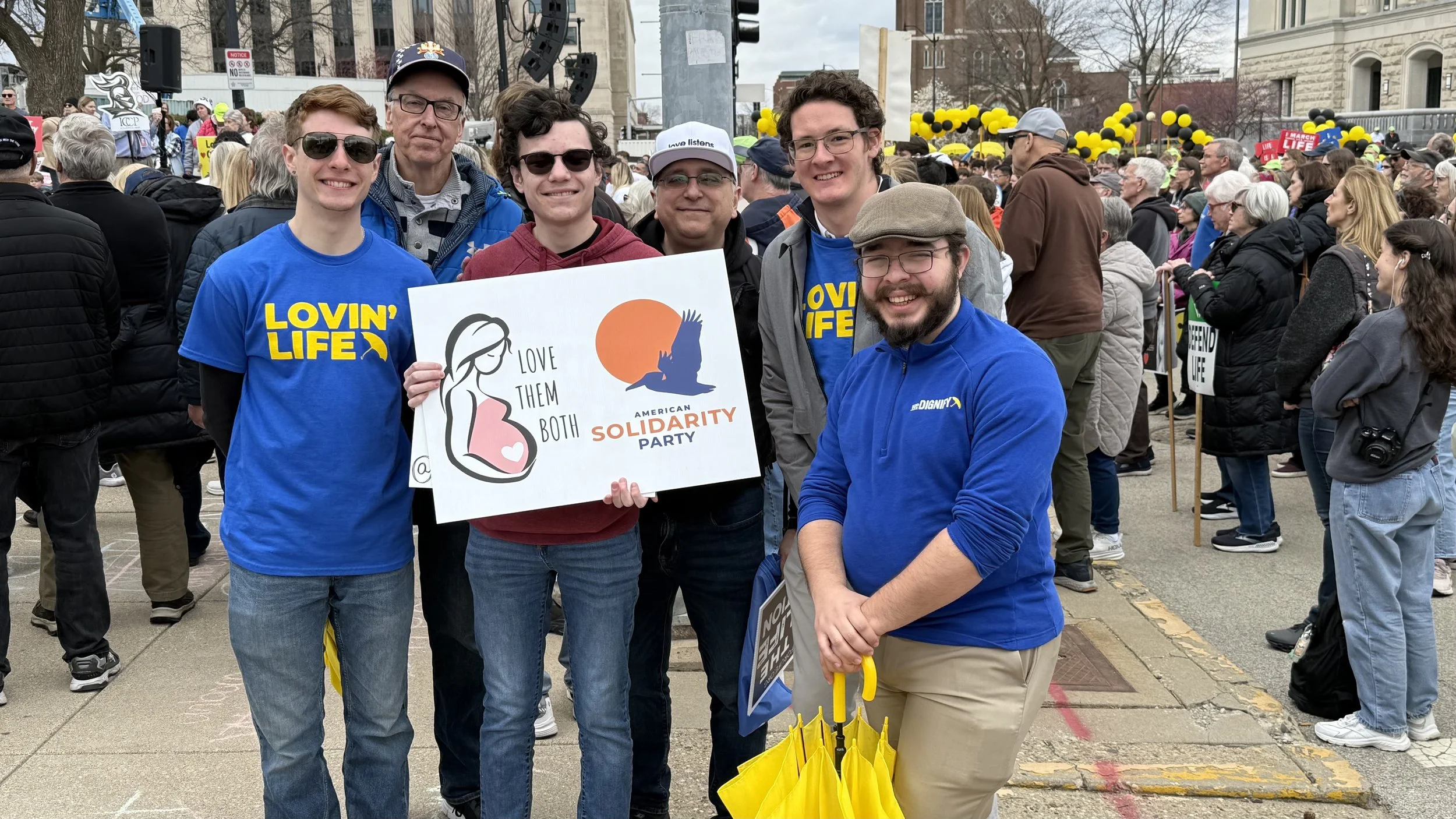 Six people standing together at a protest or rally, smiling and holding a sign that reads "Love Them Both" with a graphic of a pregnant woman and an orange sun, surrounded by a crowd of people holding yellow and black balloons and signs, in an outdoor urban setting.