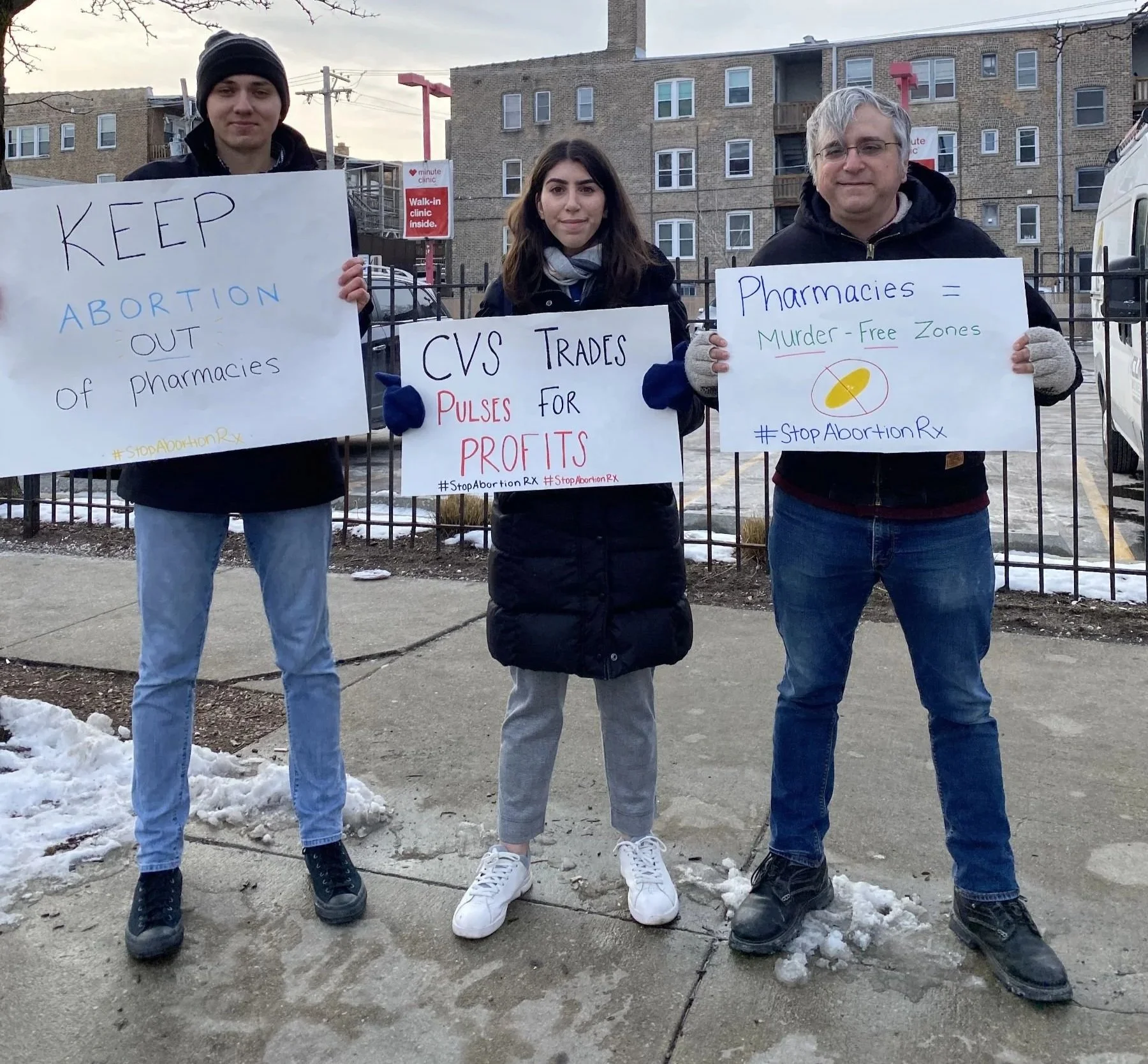 Three people standing outside on a sidewalk holding protest signs. The person on the left is a young man wearing a black beanie and black jacket, holding a sign that says 'Keep Abortion Out of Pharmacies.' The person in the middle is a young woman with long dark hair, wearing a black coat and gray pants, holding a sign that says 'CVS Trades Pulses for Profits.' The person on the right is an older man with white hair and glasses, wearing a black hoodie and gloves, holding a sign that reads 'Pharmacies = Murder-Free Zones' with a yellow capsule pill illustration. There is snow on the ground and a building with multiple floors and balconies in the background.