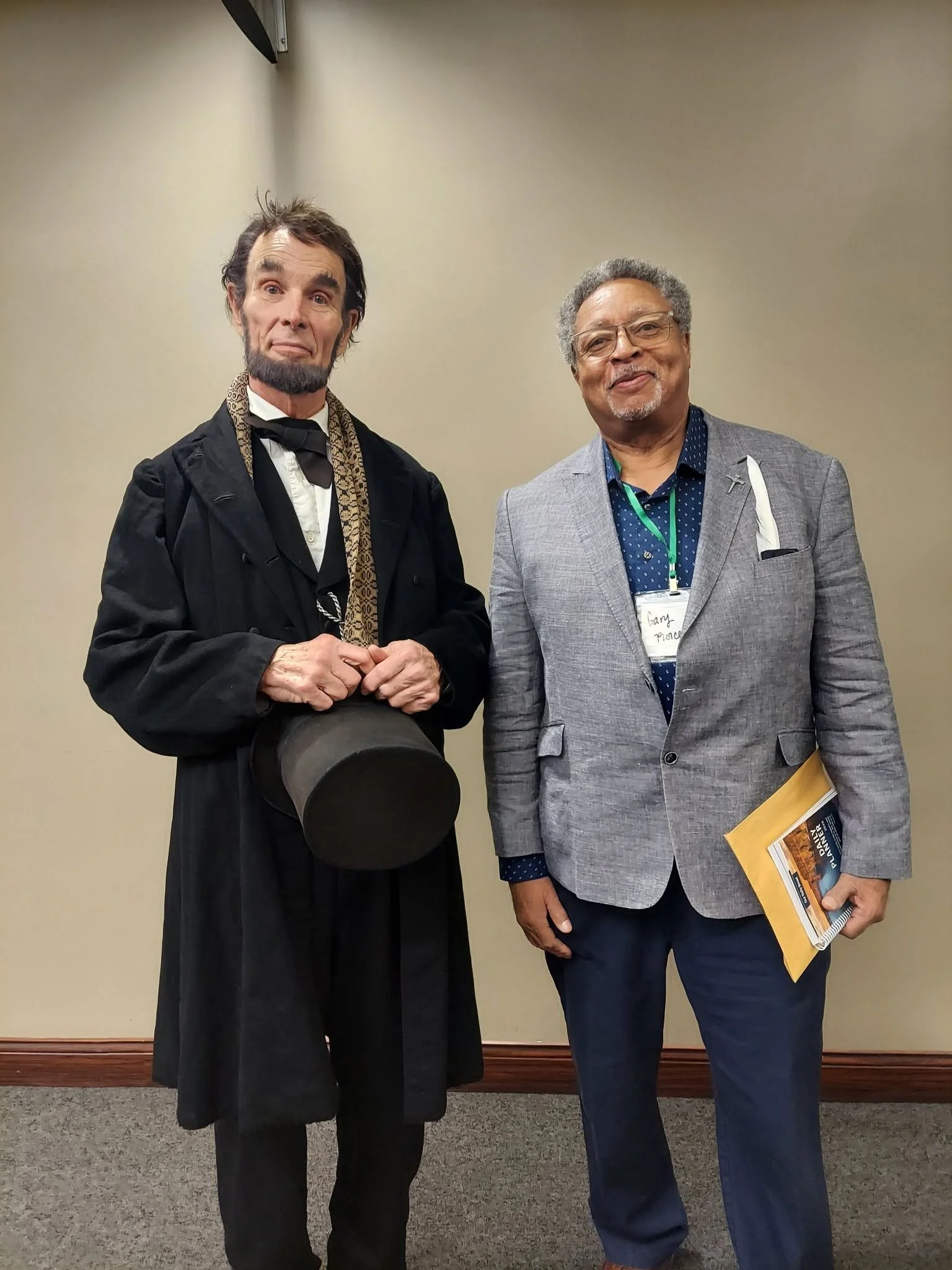 Two men standing indoors, dressed in business attire, posing for the photo. The man on the left has dark, slightly messy hair, a beard, and wears a black coat with a bow tie and scarf. The man on the right has gray hair, glasses, and is dressed in a light gray blazer with a blue shirt and dark pants, holding a yellow pamphlet and a notebook.