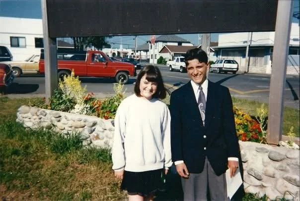 A young girl and a young man are standing outside in front of a stone-edged flower bed with yellow and red flowers. The girl is wearing a white sweater and dark skirt, while the young man is dressed in a dark blazer, white shirt, and tie. They are smiling at the camera. Buildings and parked cars are visible in the background.