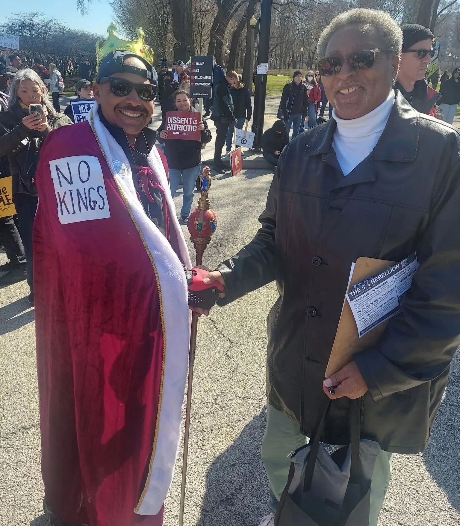 Two men shaking hands at a protest or rally. The man on the left is wearing a crown, sunglasses, a red cape, and a white shirt with a sign that says "NO KINGS". The man on the right is wearing sunglasses, a black leather jacket, and is holding a clipboard and a flyer. In the background, there are several people holding signs and banners.