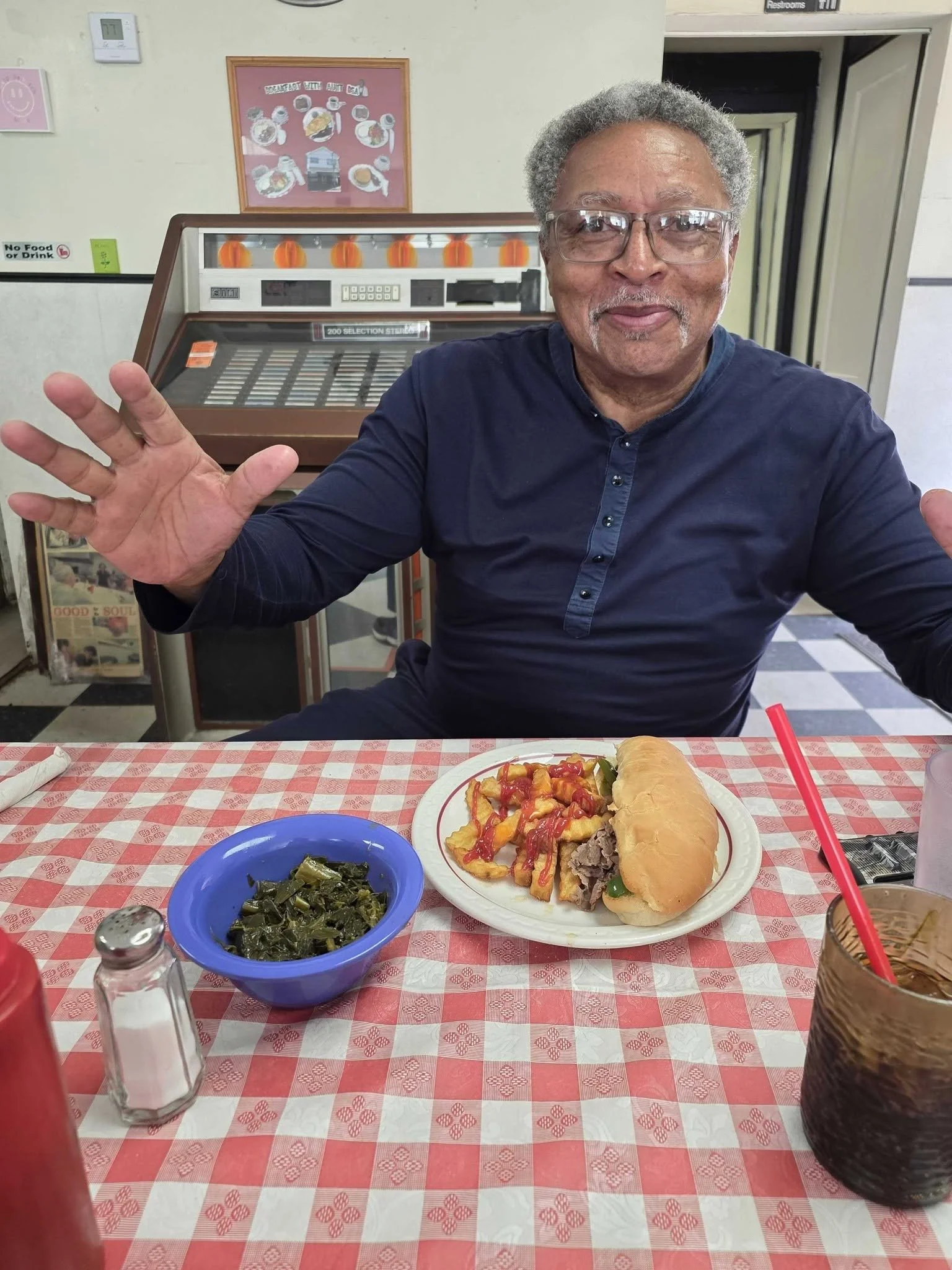 A man smiling at a restaurant table with a plate of chopped beef topped with green peppers, fried onion rings with ketchup, and a long bread roll. In front of him, a blue bowl of cooked greens and a salt shaker are visible. There is a glass of soda with a red straw, and a pitcher of iced tea. The background features an ice cream freezer, a framed picture, and a wall with various signs.