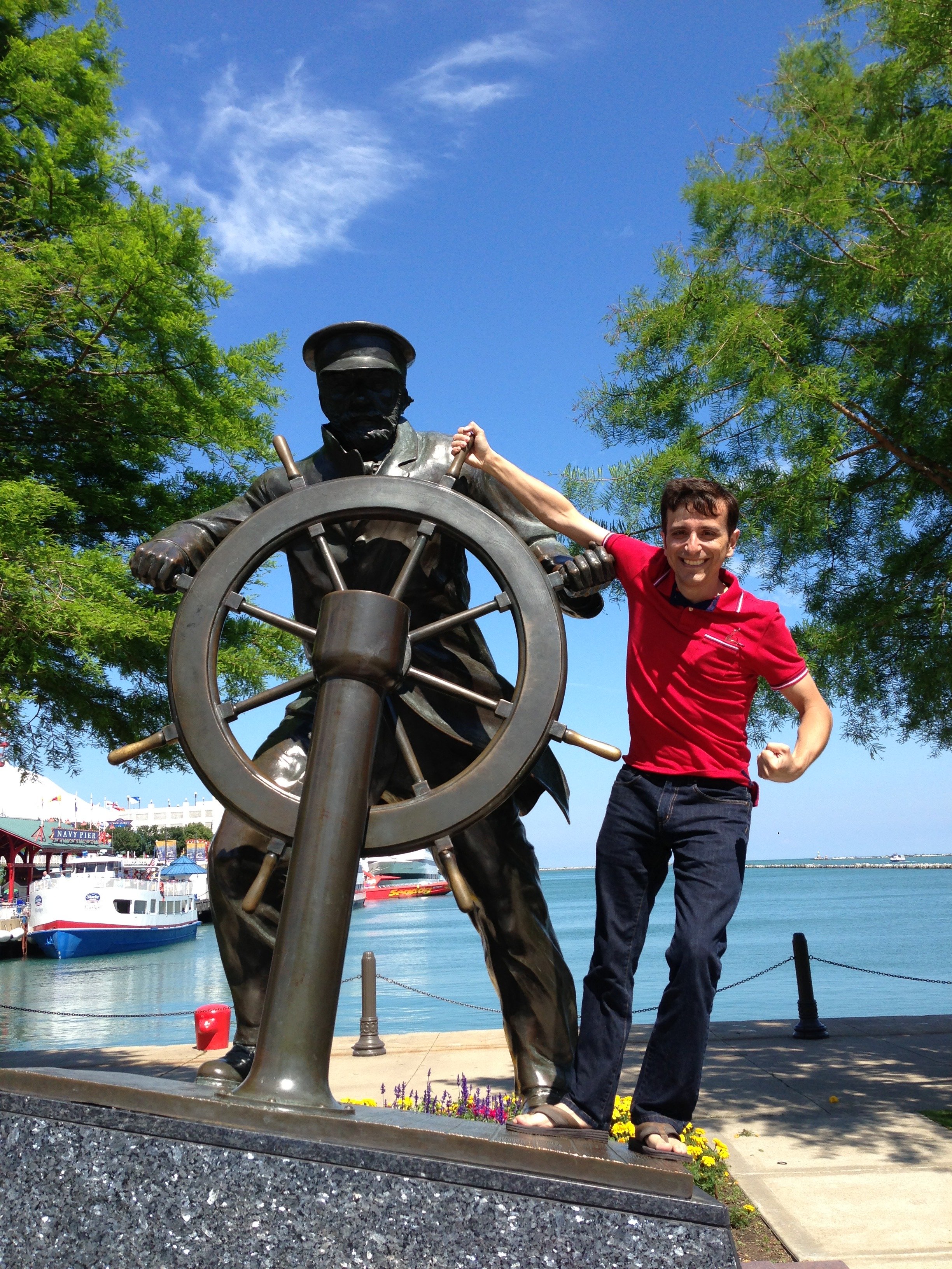 A young man in a red shirt and jeans standing on a concrete platform near a harbor, smiling and posing with a bronze statue of a ship captain at a ship's wheel, with boats, water, and trees in the background.