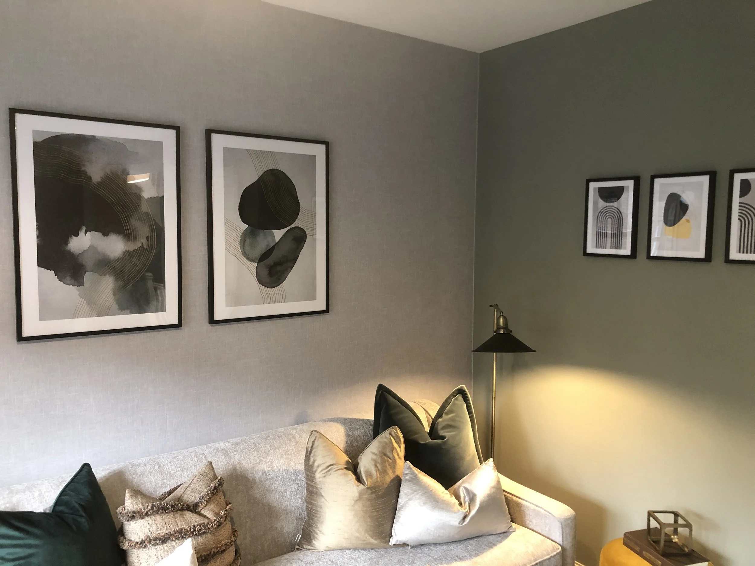 Living room corner with a beige sofa, decorative pillows, wall art, and a floor lamp, with a two-tone wall background.