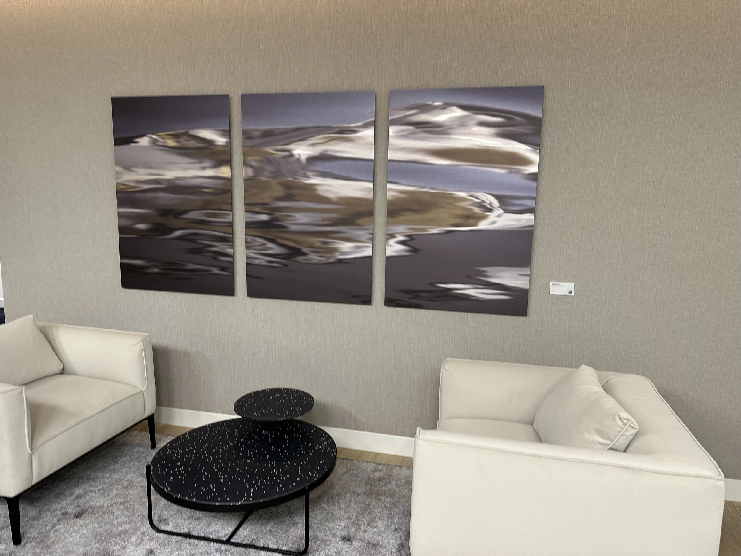 A modern living room with two white sofas, a black coffee table with small white dots, and a three-panel artwork depicting a distorted water reflection on the wall.