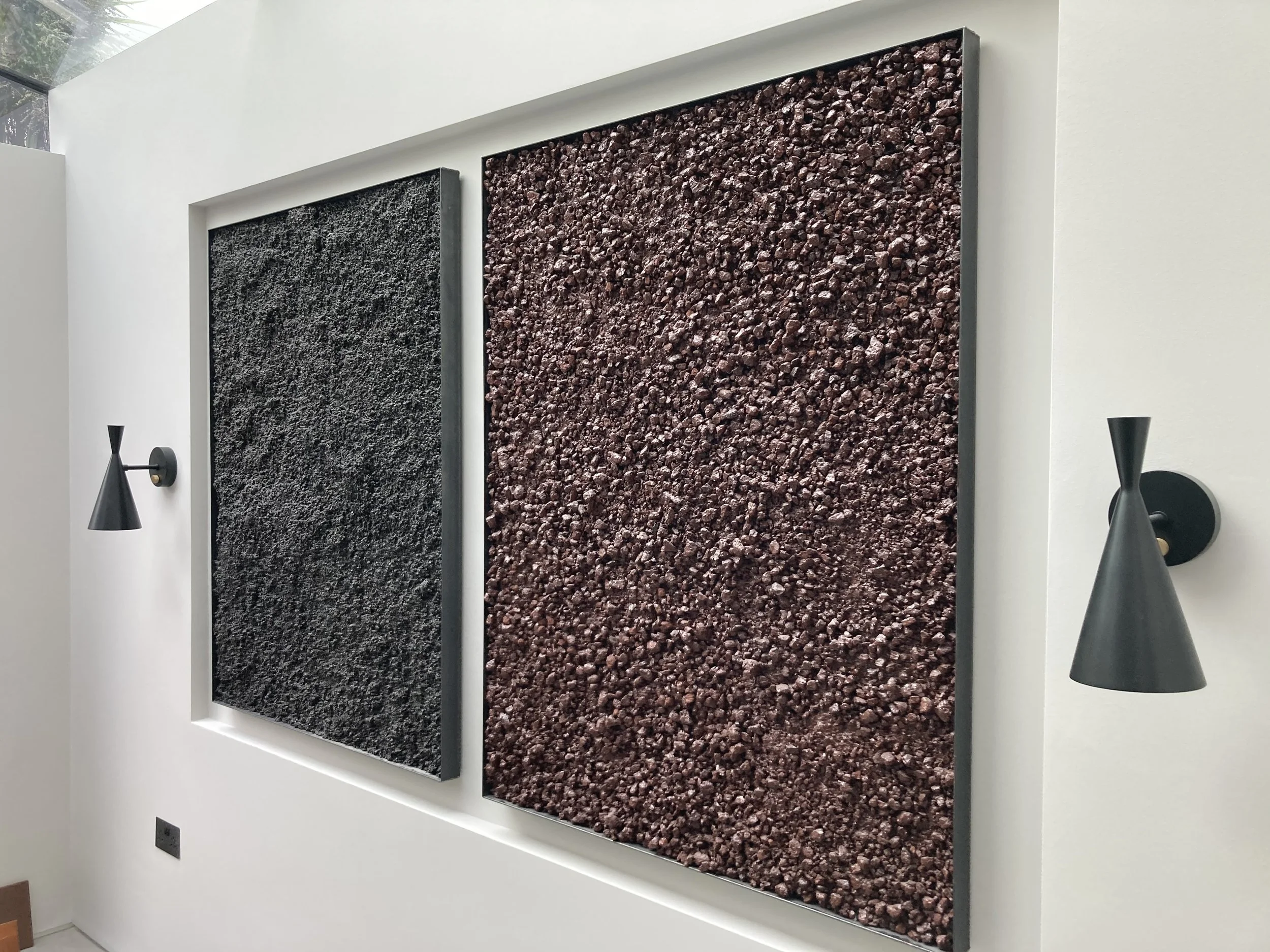 Two textured wall art panels, one dark gray and one reddish-brown, mounted on a white wall in a modern interior, with black wall sconces on either side.