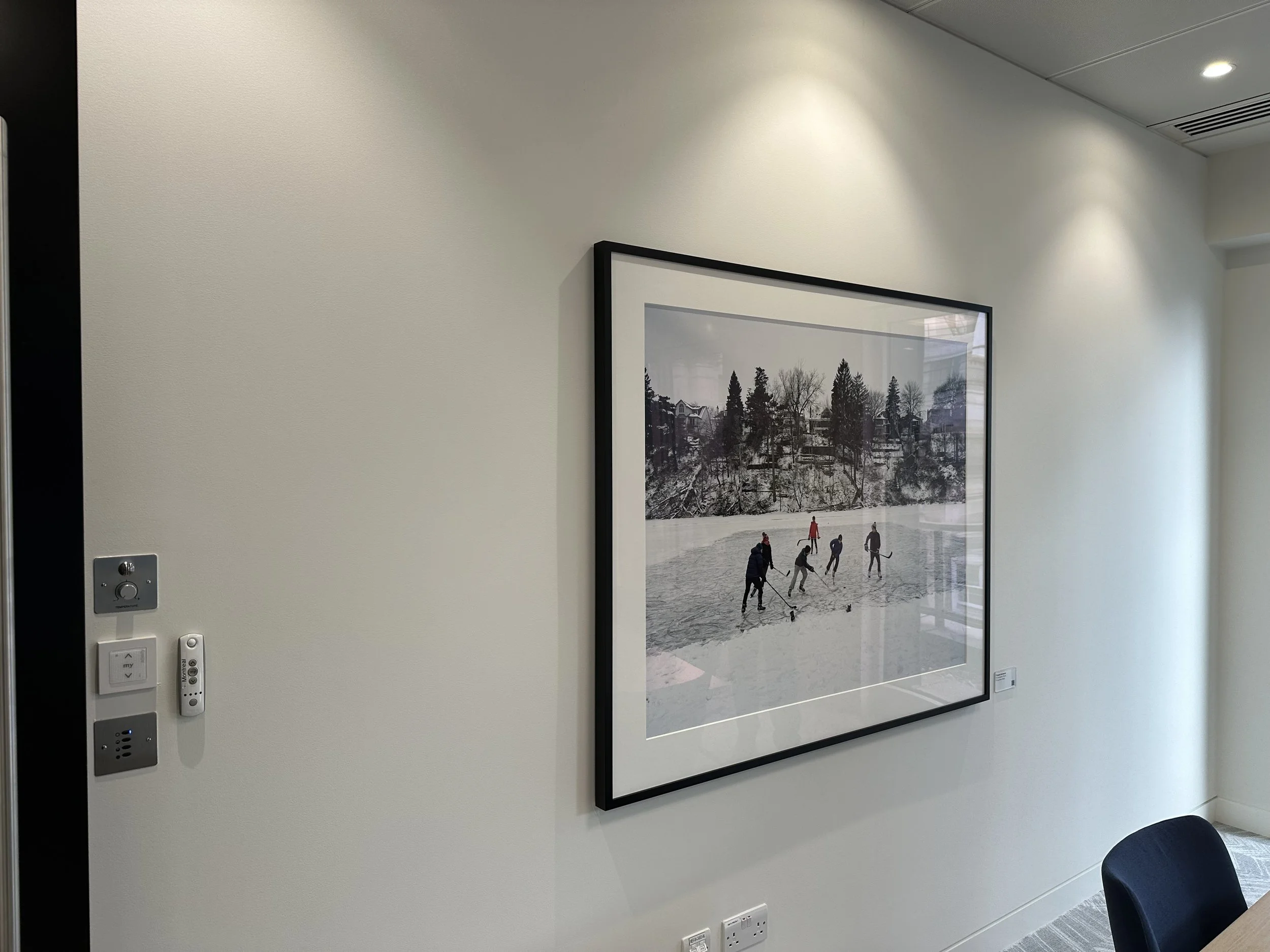 Photograph of an art print on a white wall showing a winter landscape of seven people playing hockey on a frozen pond in front of trees and houses, with reflections of the room's interior in the glass.